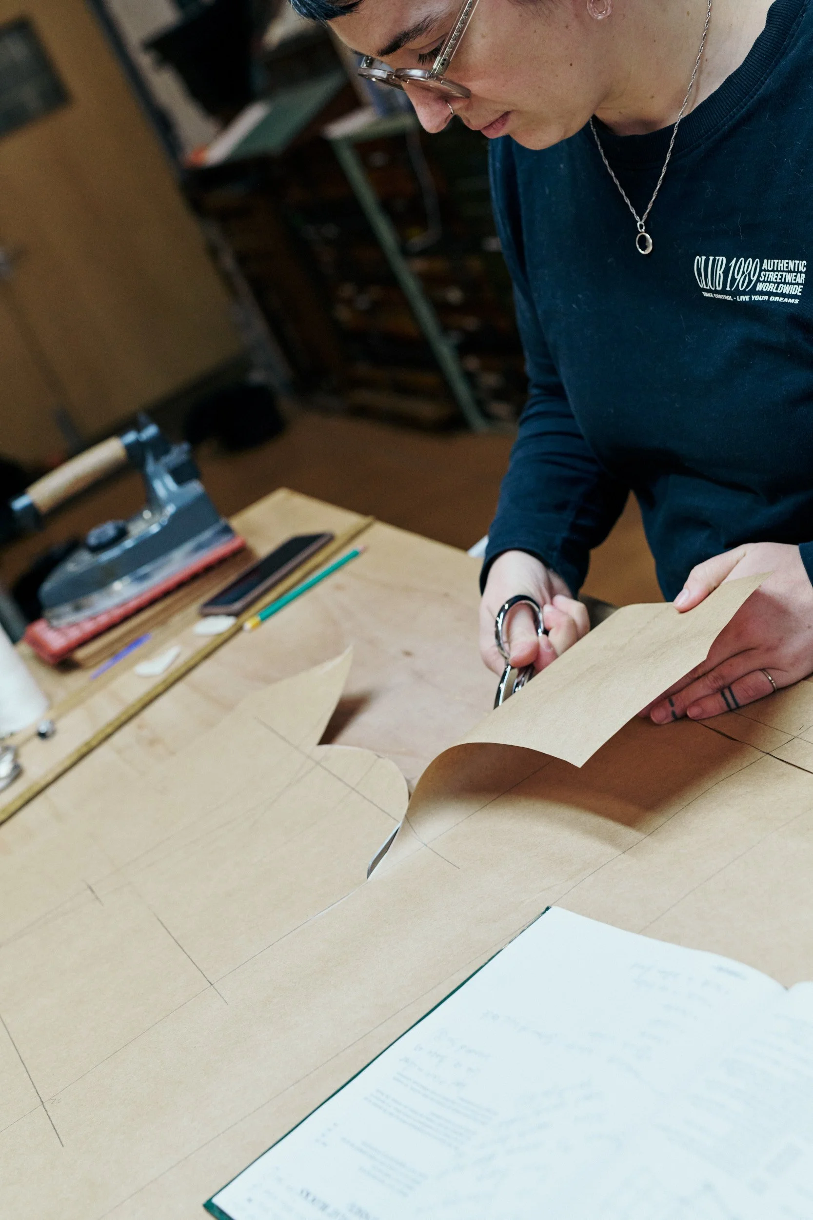 A person with glasses and rings is cutting a piece of light brown material, possibly paper or fabric, with scissors at a workbench. The workbench has tools and rulers on it, and a notebook with handwritten notes is visible in the foreground.