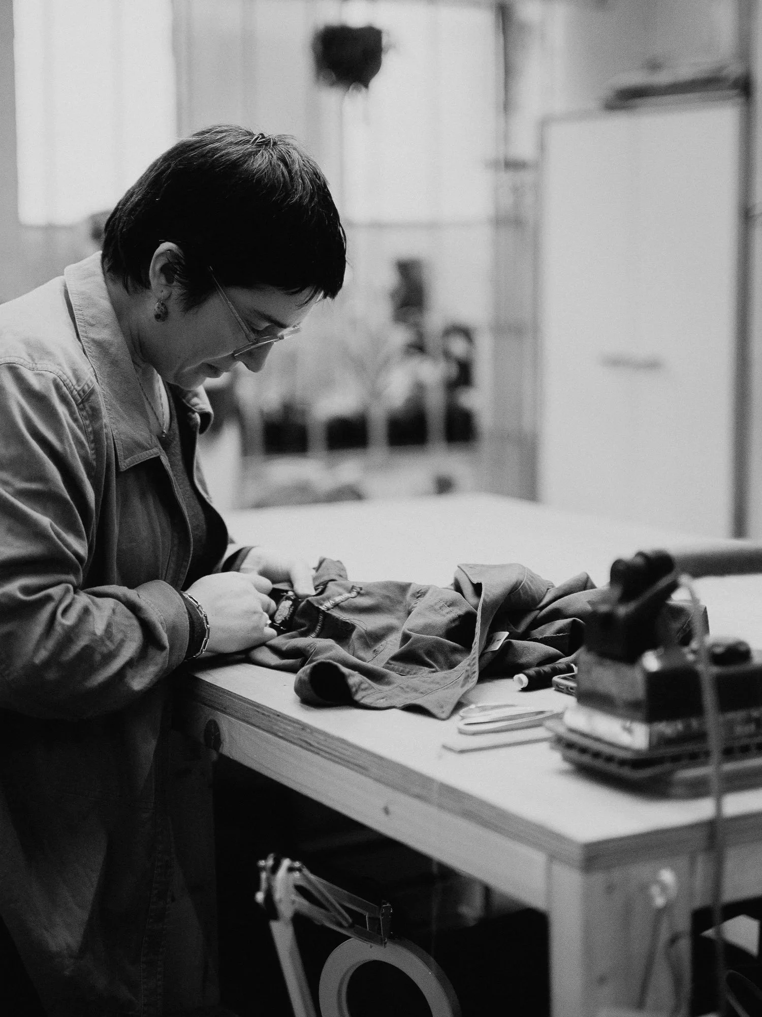 A woman with short hair and glasses working on a sewing project at a wooden workbench, with sewing tools and fabric laid out on the table.