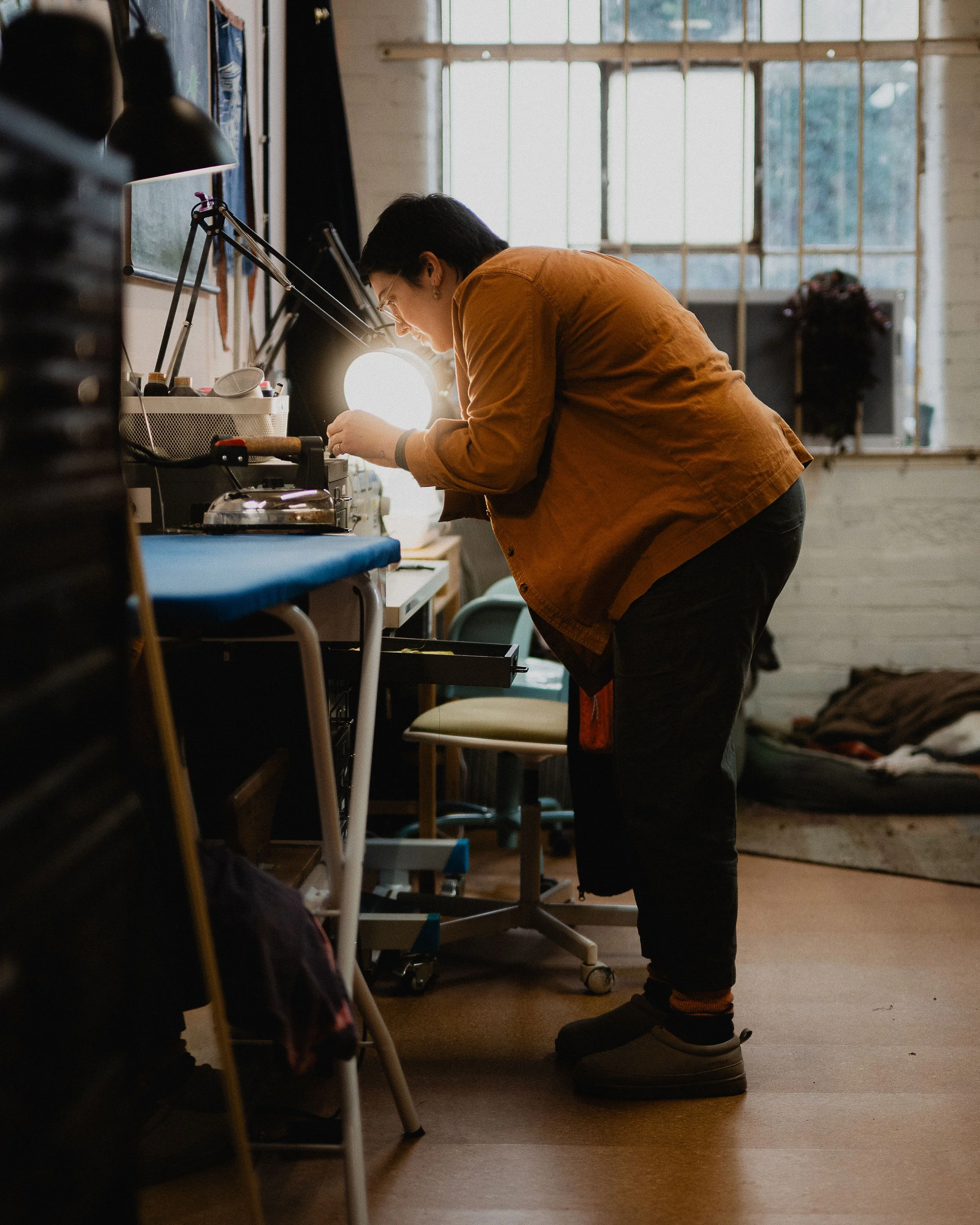 Person leaning over a workspace, working on a project in a well-lit room with large windows, cluttered with tools, a lamp, and various objects.