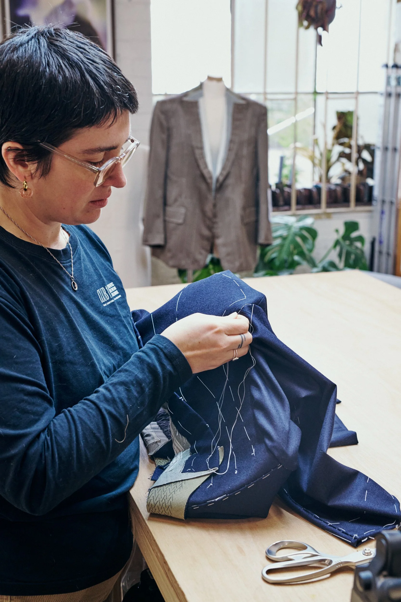 A woman sewing navy blue fabric with white thread in a workshop. A tailored suit jacket on a mannequin and plants in the background.