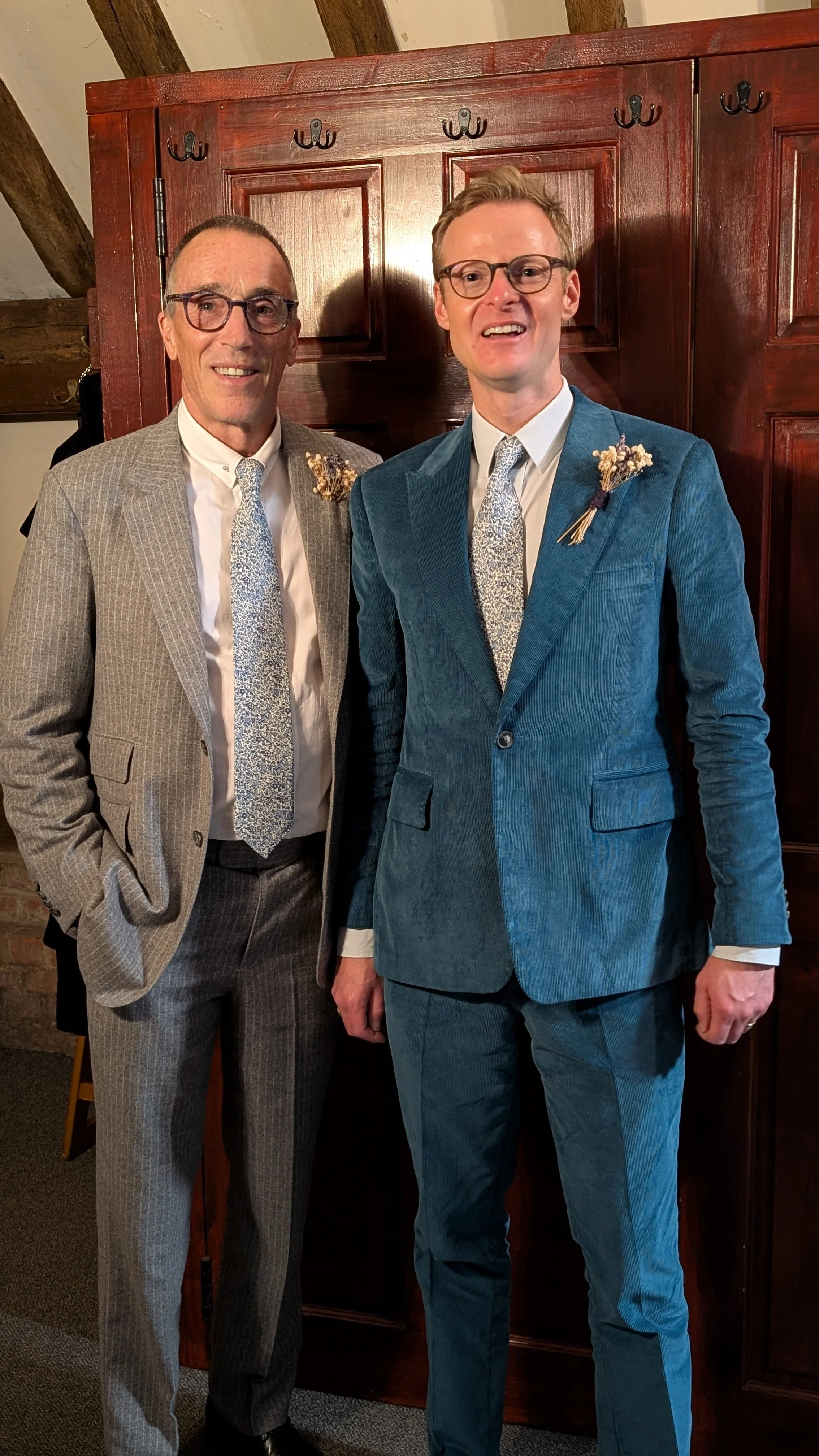 Two men dressed in suits with floral boutonnières, standing indoors in front of a wooden cabinet and exposed beams.