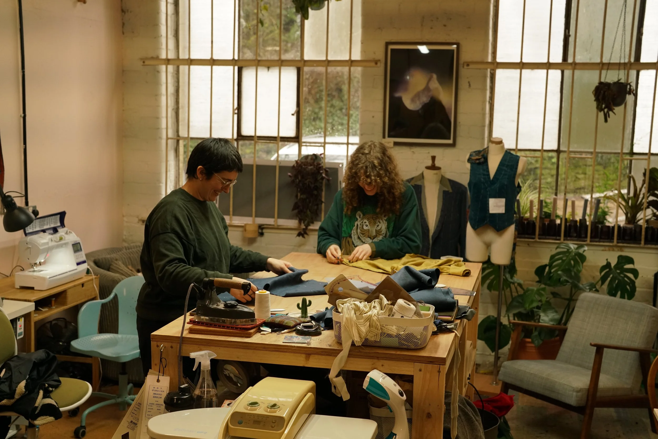Two women working at a sewing table in a creative studio surrounded by fabrics, mannequins, and plants.
