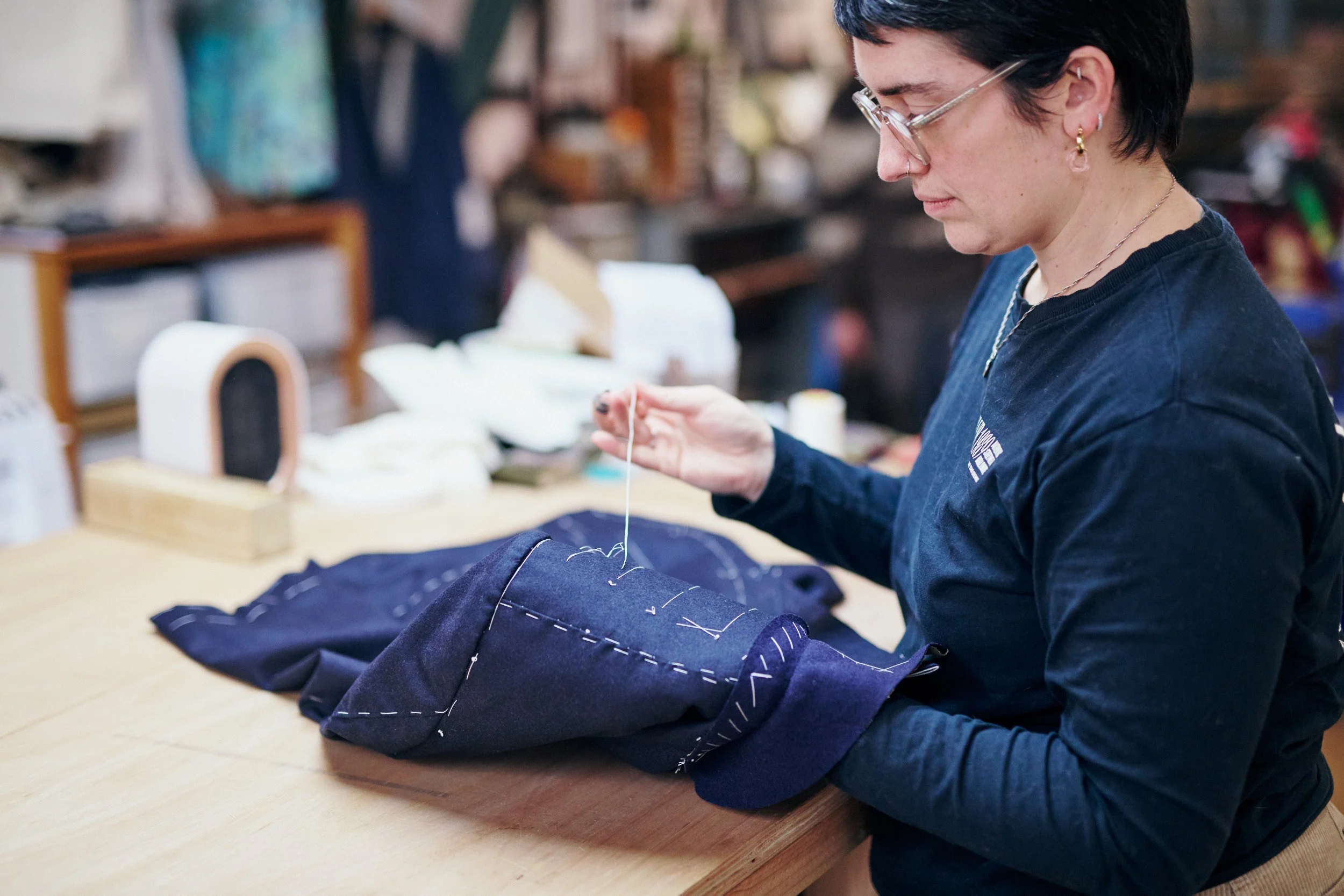 A woman sewing navy blue fabric on a wooden table in a workshop environment.