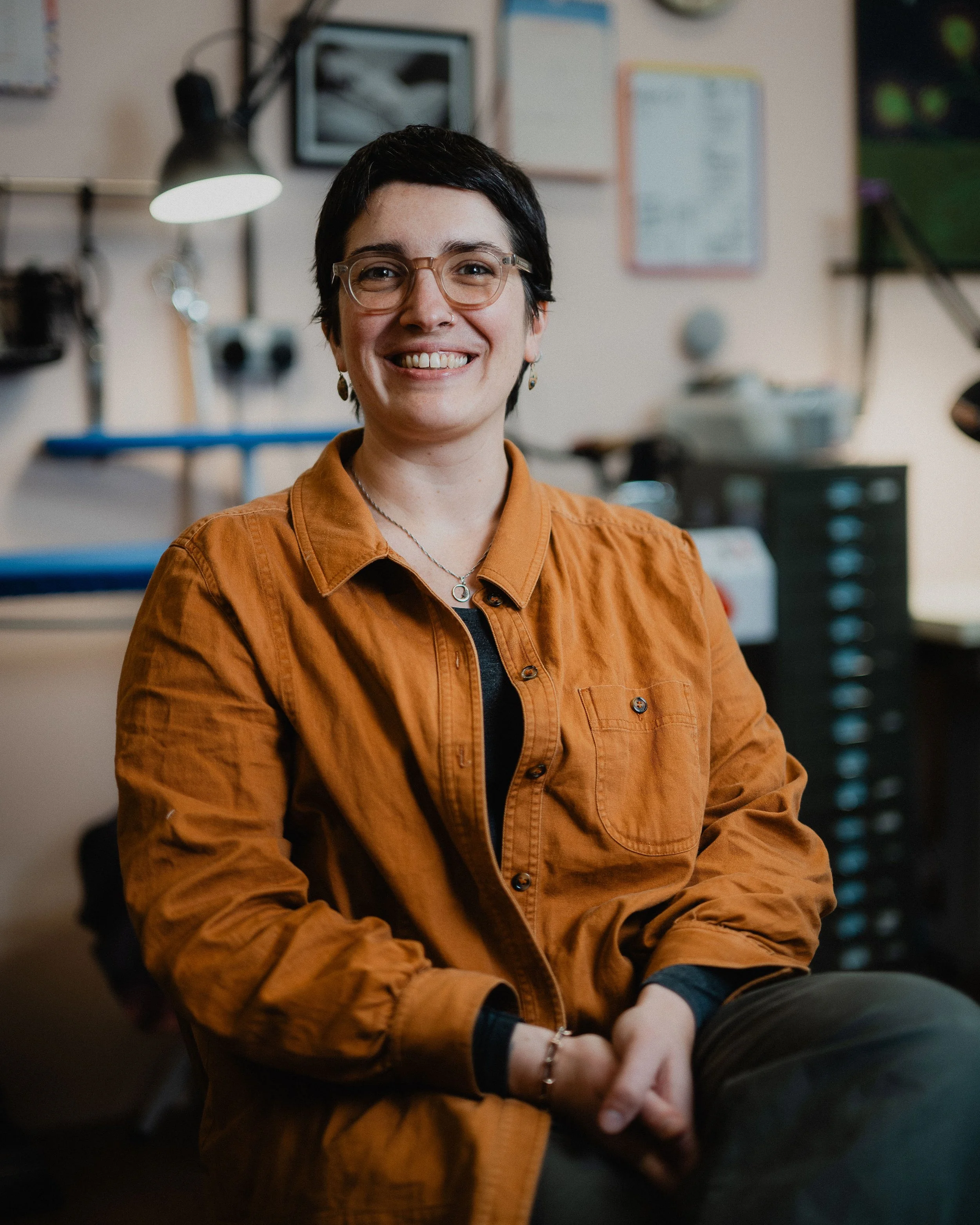 A woman with short dark hair, glasses, and a nose ring smiling at the camera in an indoor workspace, wearing an orange-brown jacket and jewelry.