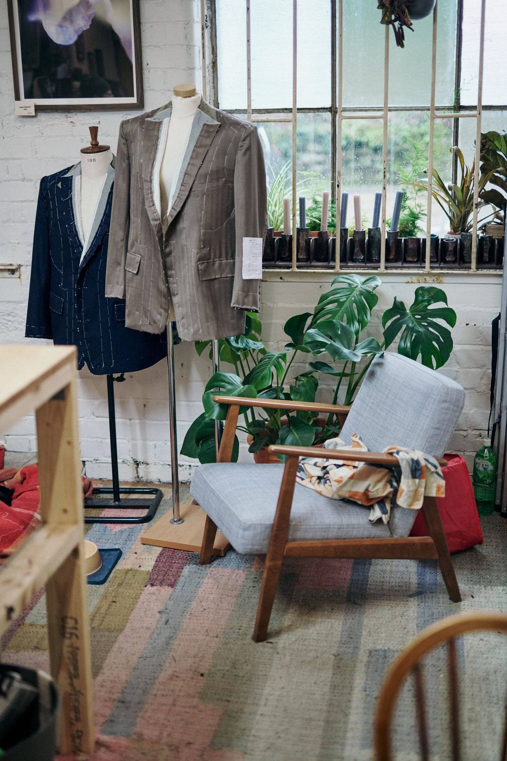 Interior of a boutique or studio featuring two mannequin suits, a gray armchair with a scarf, green plants, candles in pots on window sill, and a colorful rug on the floor.