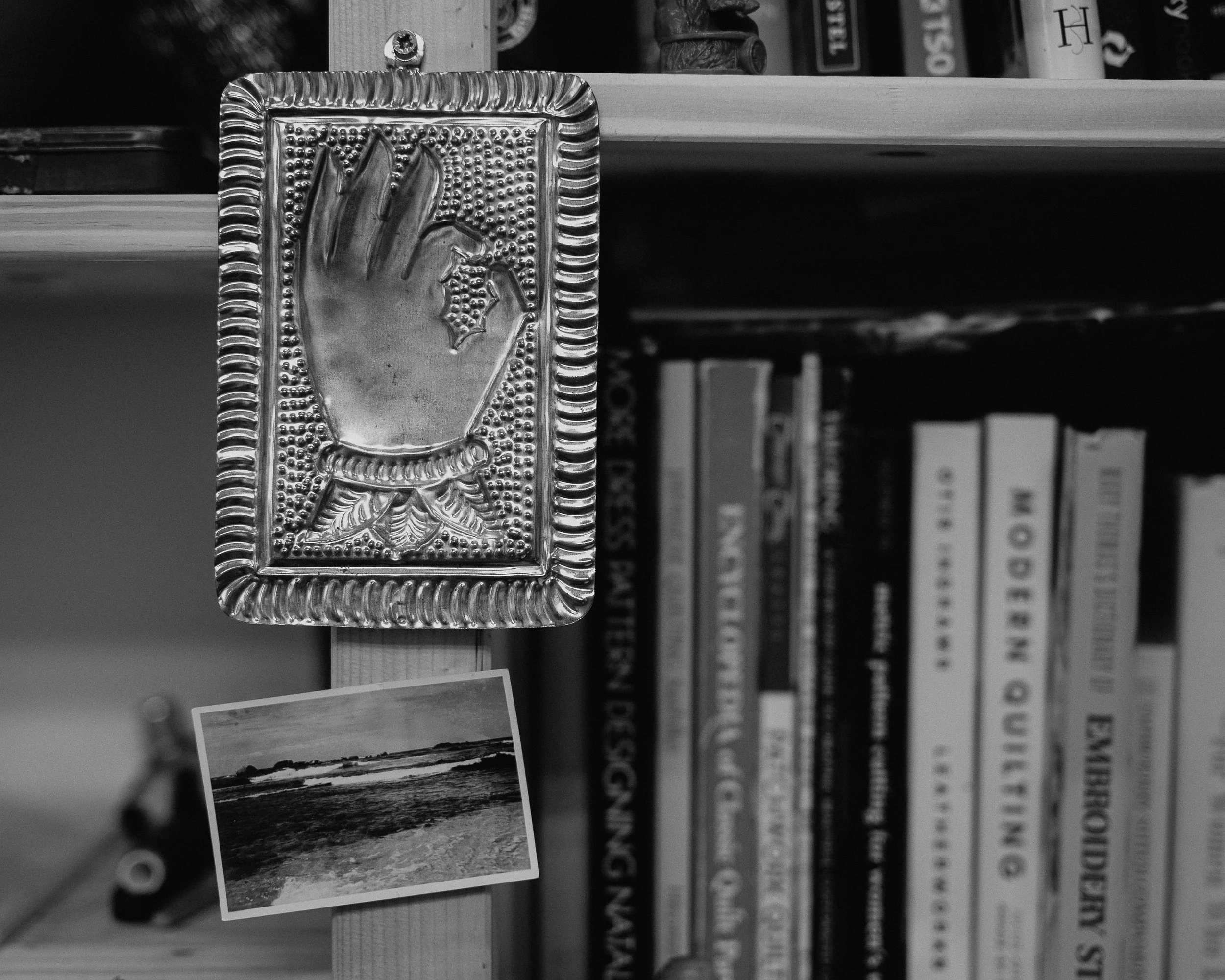 Black and white photo of a bookshelf with a silver religious icon and a small photograph of a beach scene attached to a wooden support.