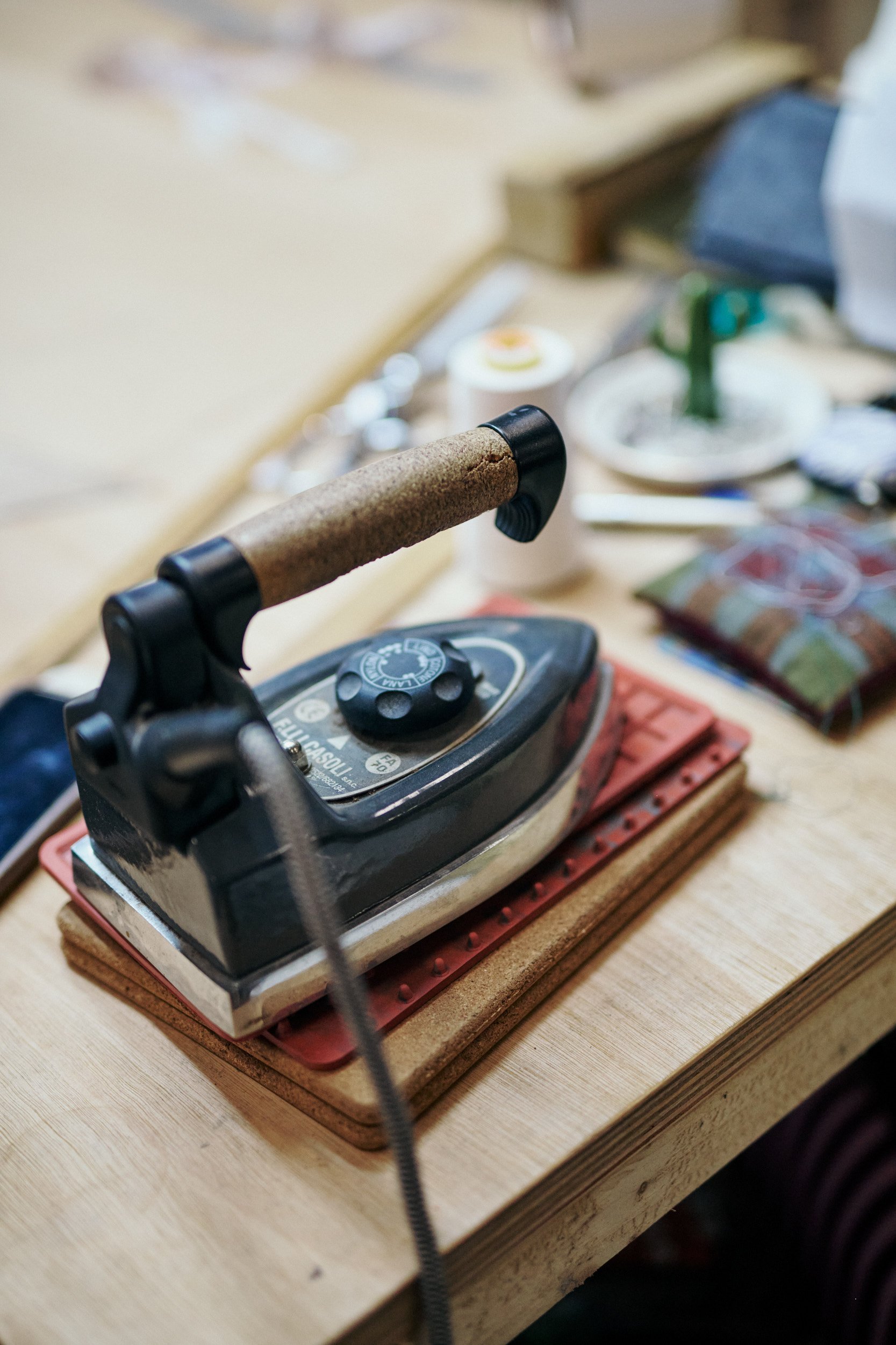 An electric iron resting on a cork pad on a wooden table, with various craft supplies and objects blurred in the background.