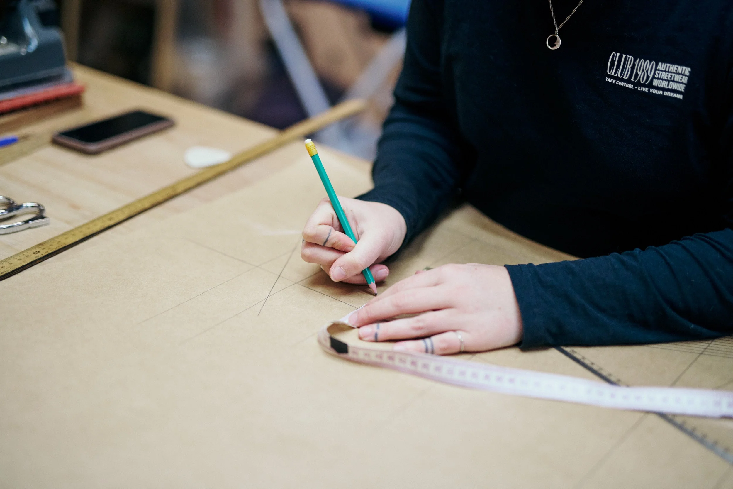 Person drawing measurement lines on a large piece of brown paper with a pen, surrounded by a measuring tape, smartphone, and scissors on the table.