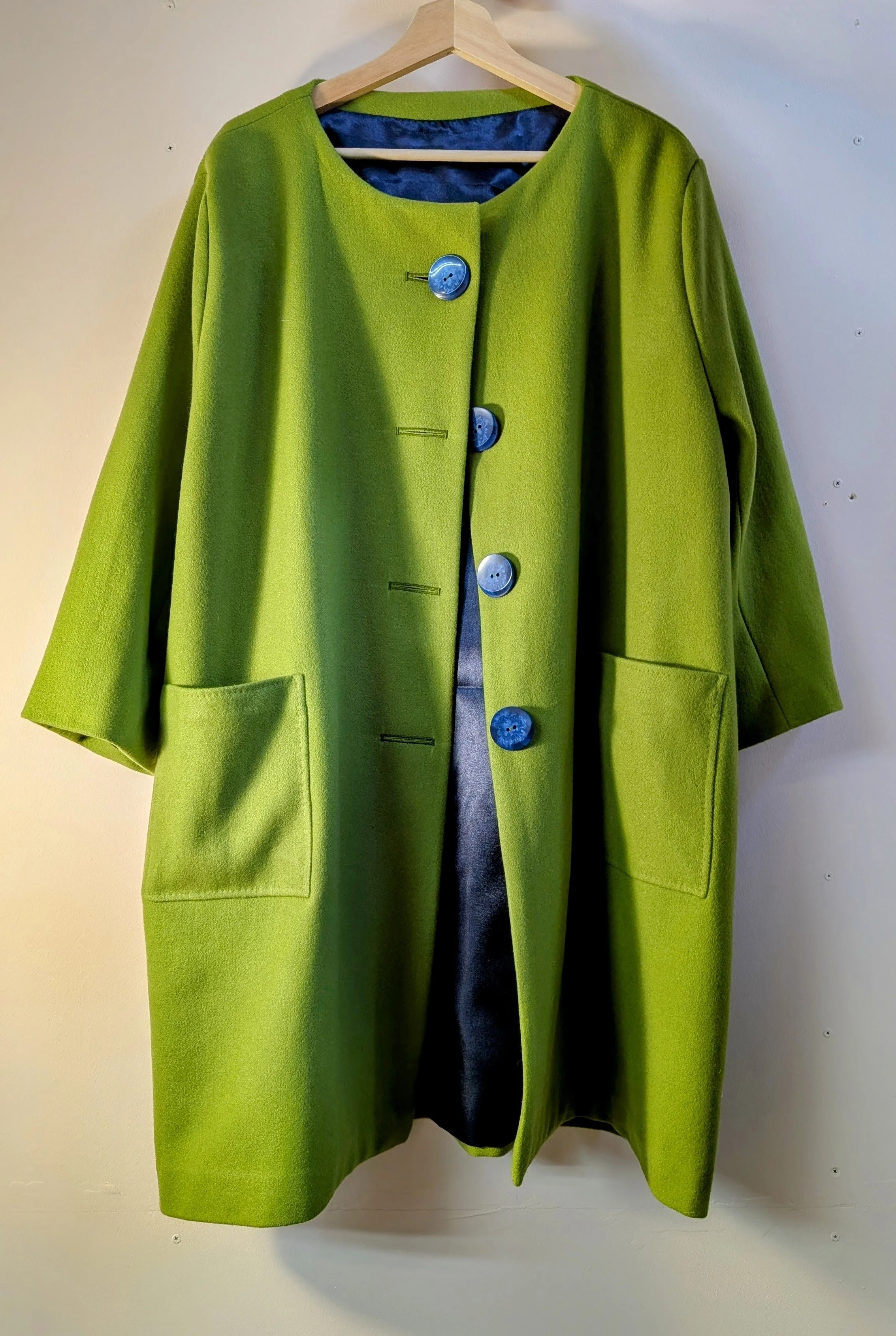 A bright green long coat hanging on a wooden hanger against a white wall. The coat has large pockets and four blue buttons.