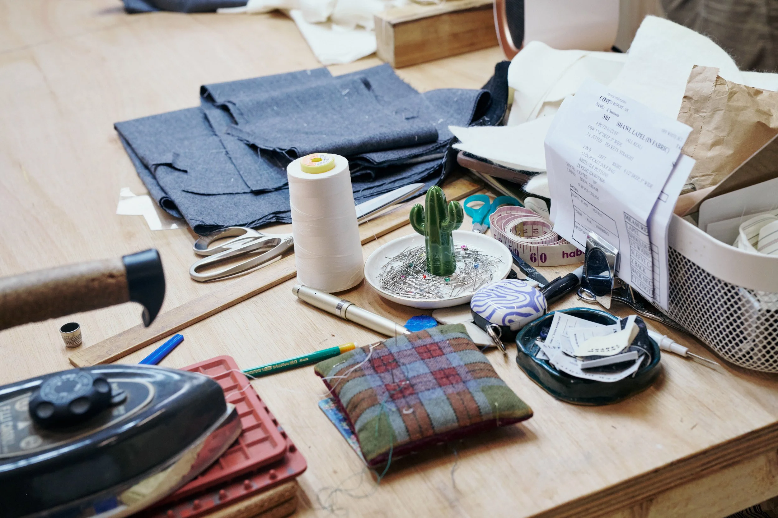 Cluttered sewing table with fabric, scissors, sewing tools, tape measure, spool of thread, pincushion, and pattern papers.