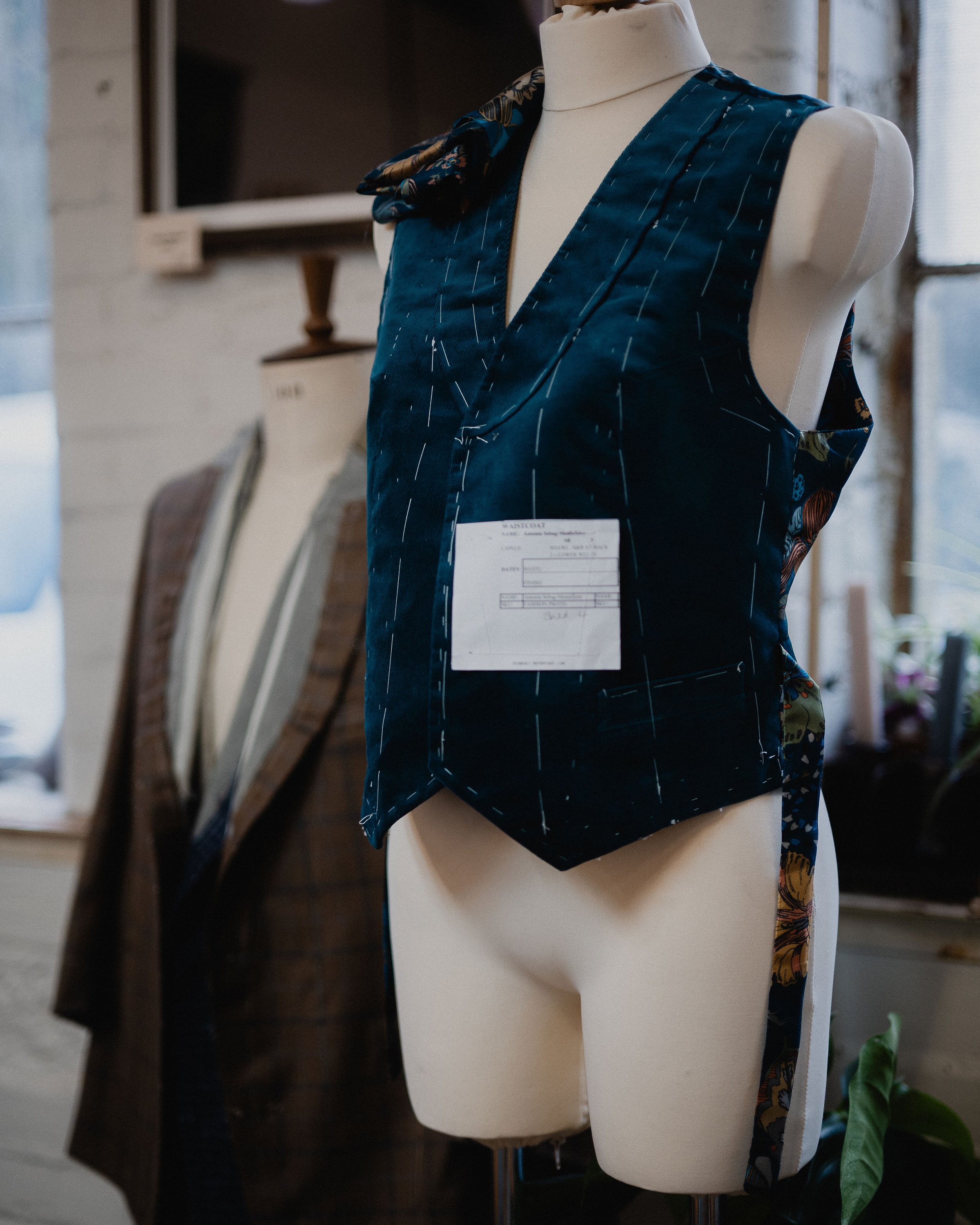 A dress form wearing a dark blue vest with a floral pattern and tag attached, in a room with fabric samples and a window.