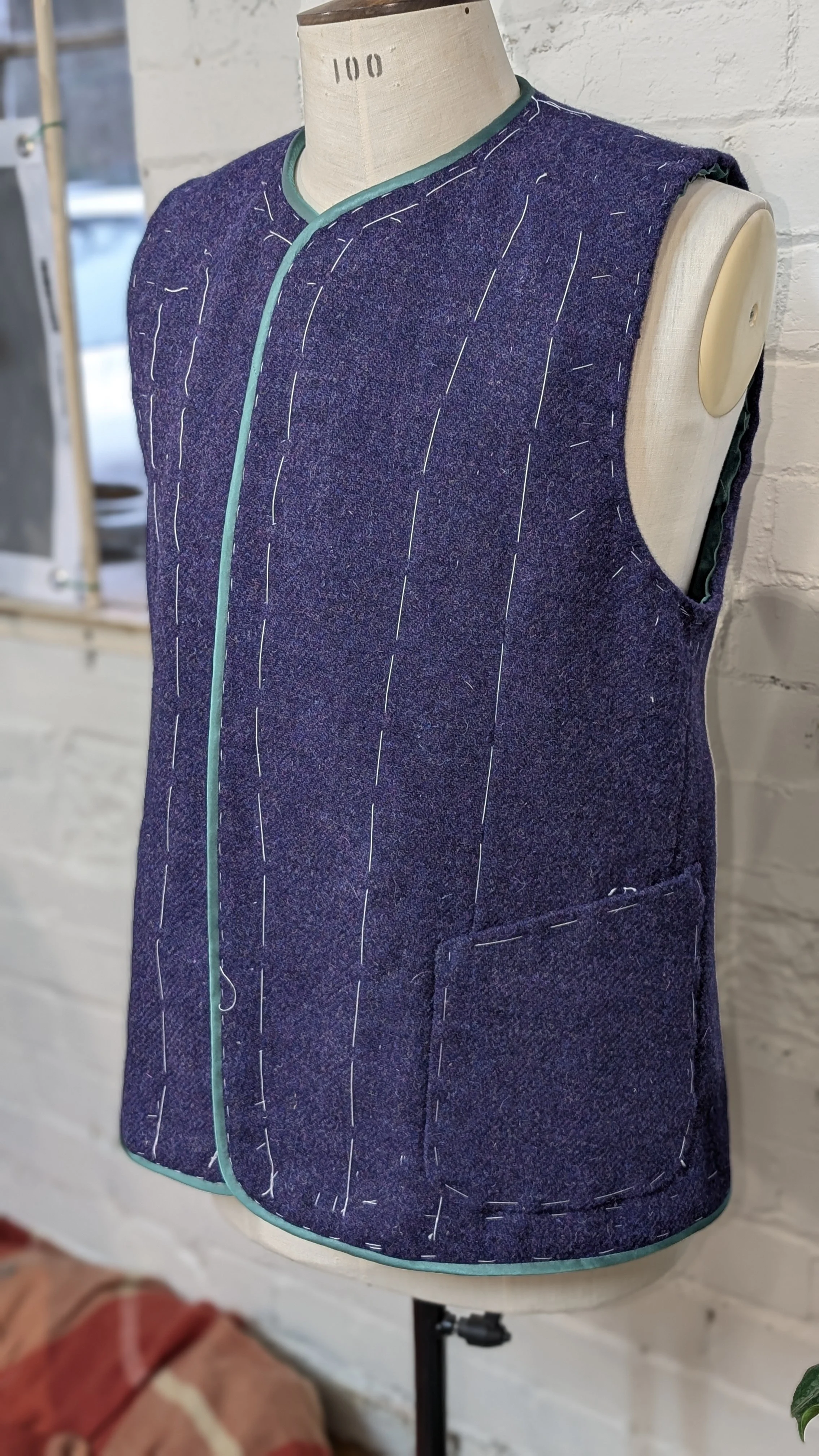 A purple sleeveless sewing vest on a dress form with white, blue, and green details, positioned against a white brick wall.