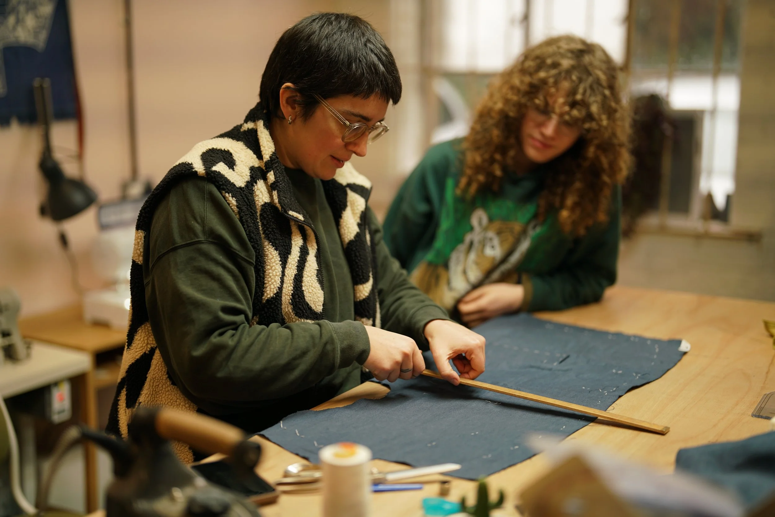 Two women working on a craft project at a wooden table, one measuring fabric with a wooden ruler, in a well-lit room with various tools and supplies.