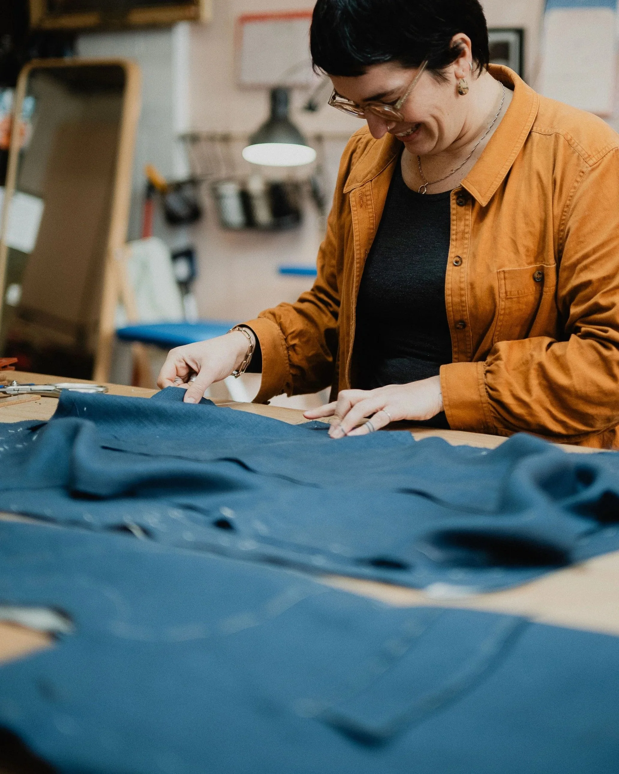 A woman is working with blue leather fabric at a workbench in a workshop, smiling and inspecting the material.