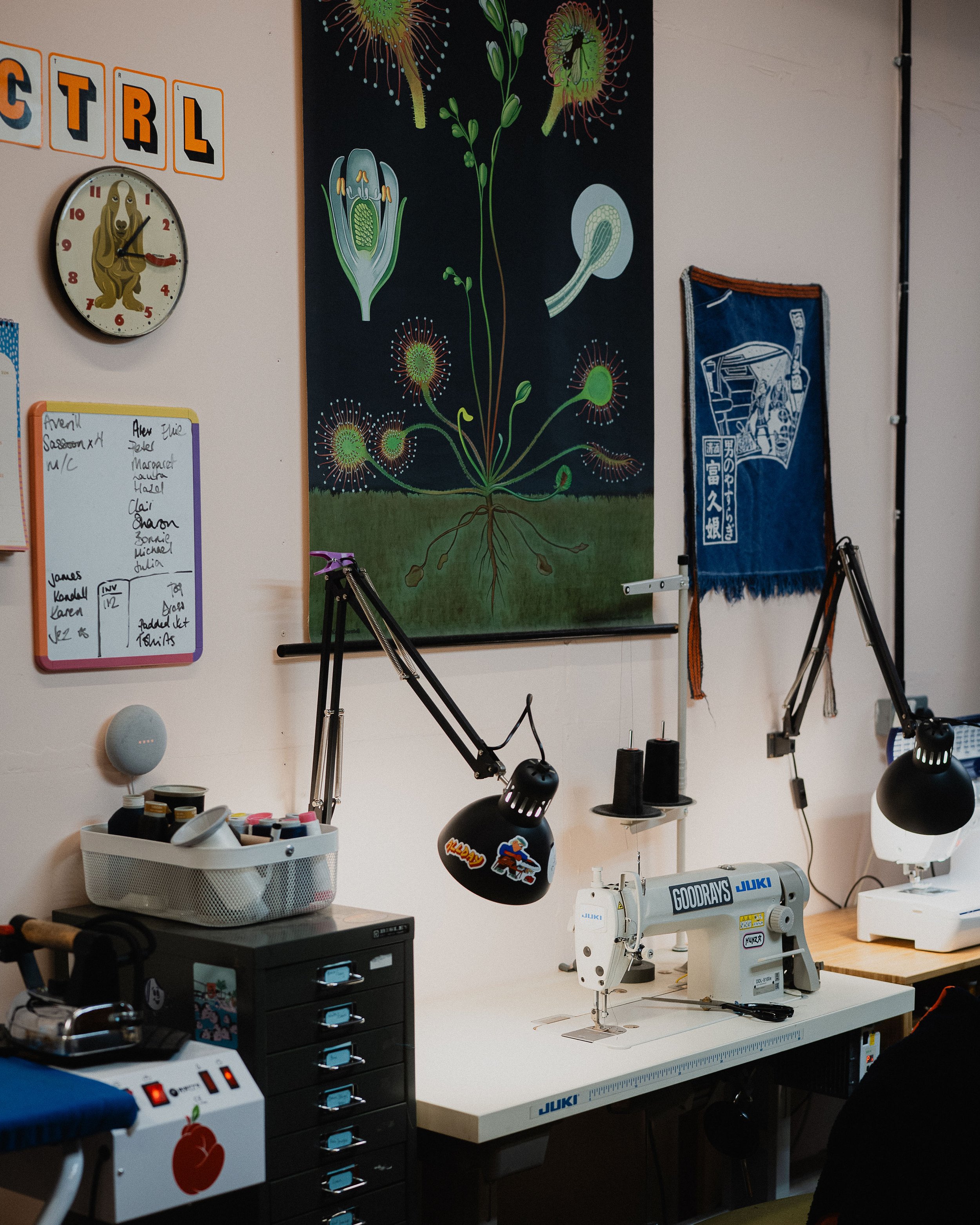 A sewing workspace with a Juki sewing machine, adjustable lamps, a whiteboard with notes, a poster of a plant anatomy, a clock with an animal design, and various tools and supplies.