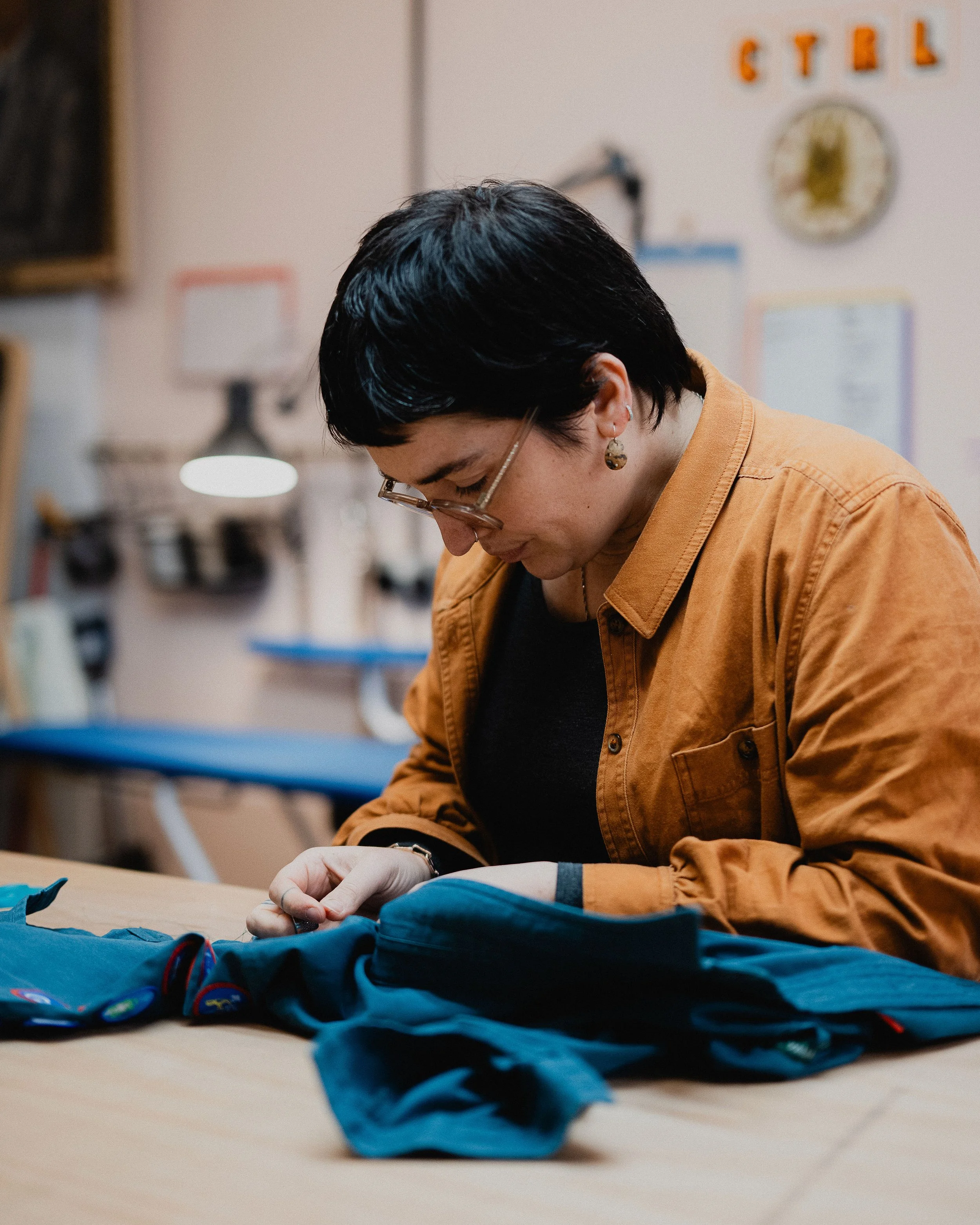 Woman in glasses sewing on a blue fabric in a workshop.