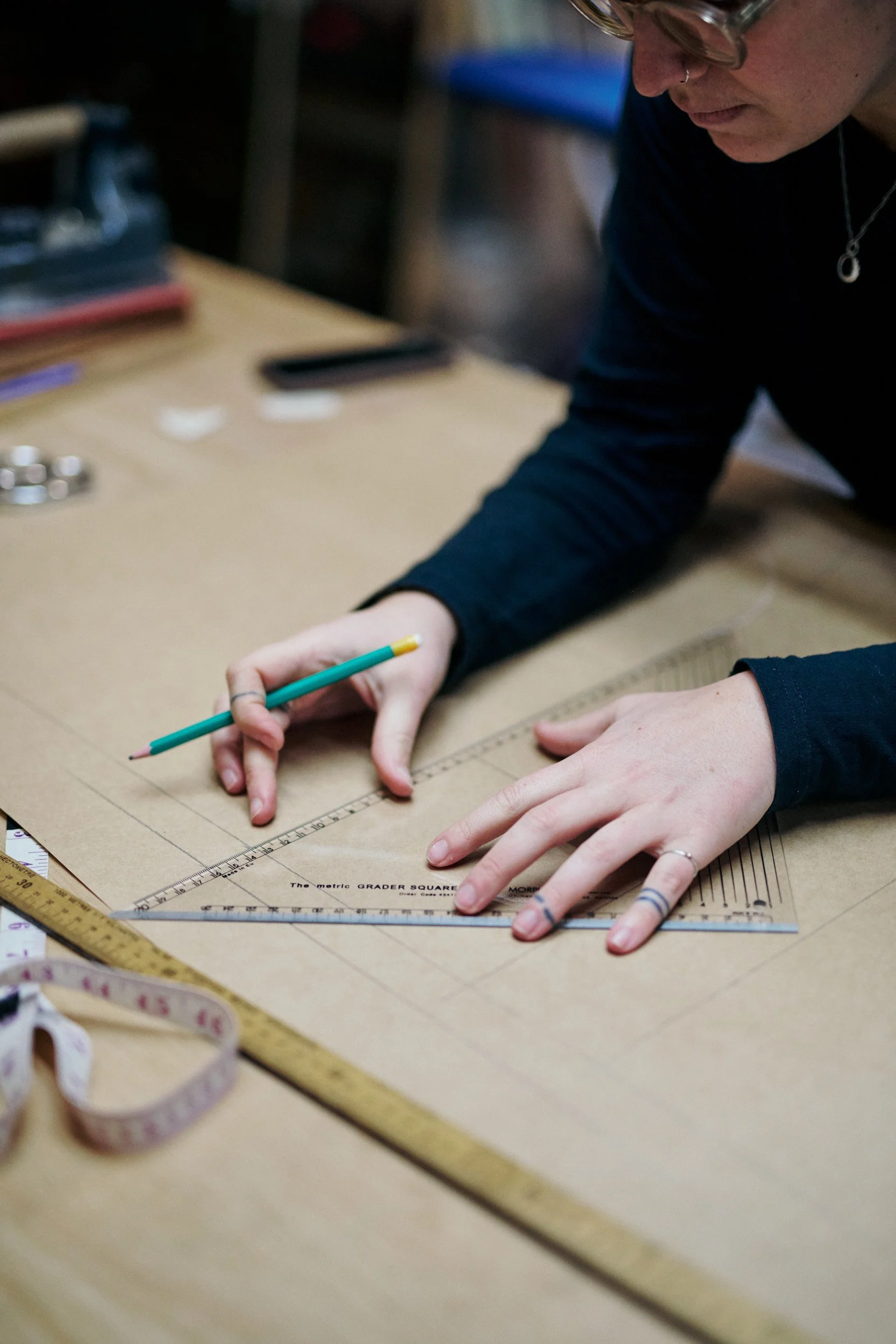 Person using a ruler and pencil on a large piece of paper for drafting or drawing at a work table, with measuring tools and supplies around.