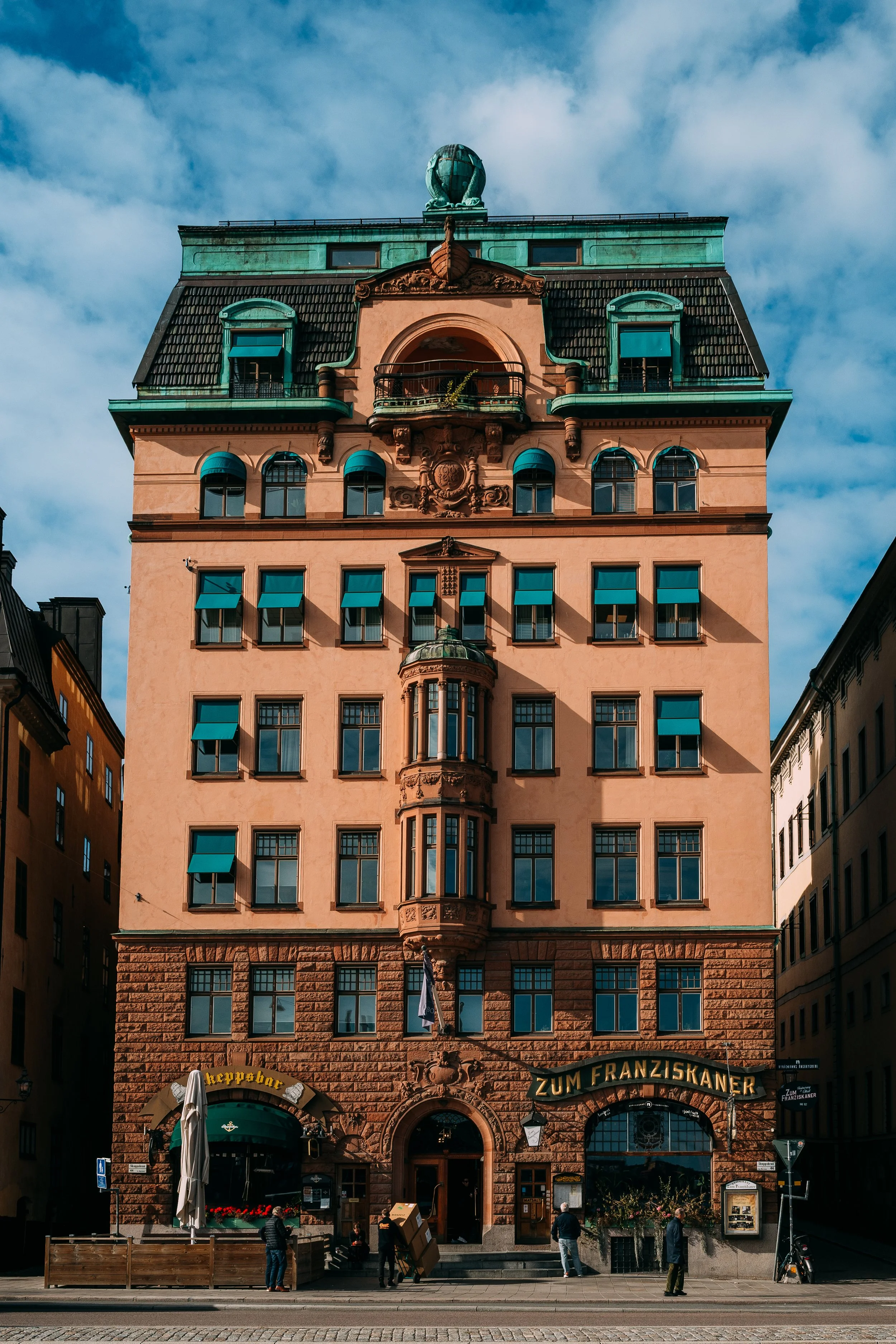 A multi-story historic building with ornate architecture, green rooftop decorations, and blue window awnings, located on a city street with people standing in front and signs reading "Zum Franziskaner" and "Kellneppner".