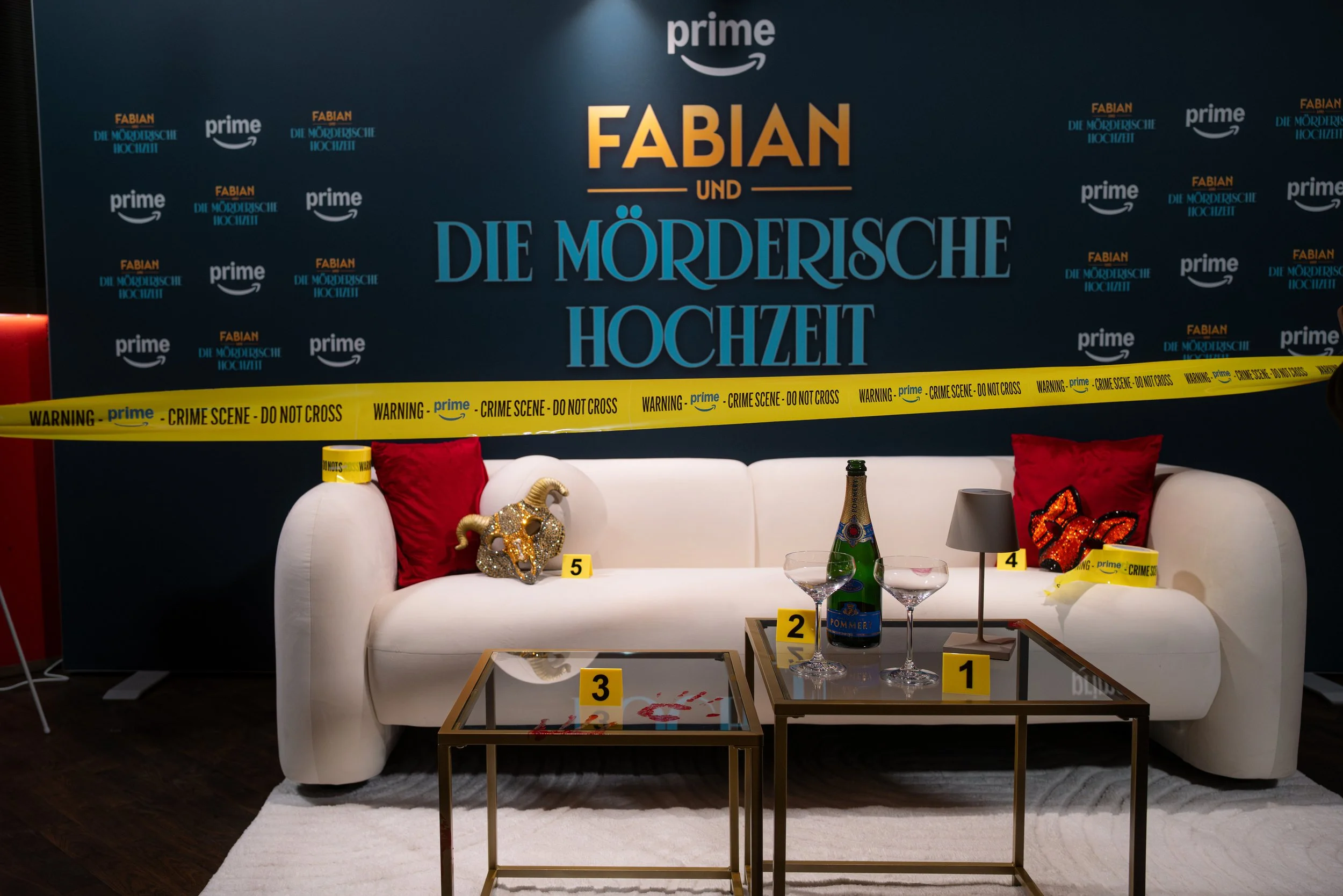 Sofa mit roten Kissen, goldener Ziegenkopf, Wein- und Wasserflaschen, Tische mit Nummern, Warnkordel, Hinweise auf eine Crime-Show. Filmpremiere Amazon, Fabian und die mörderische Hochzeit.