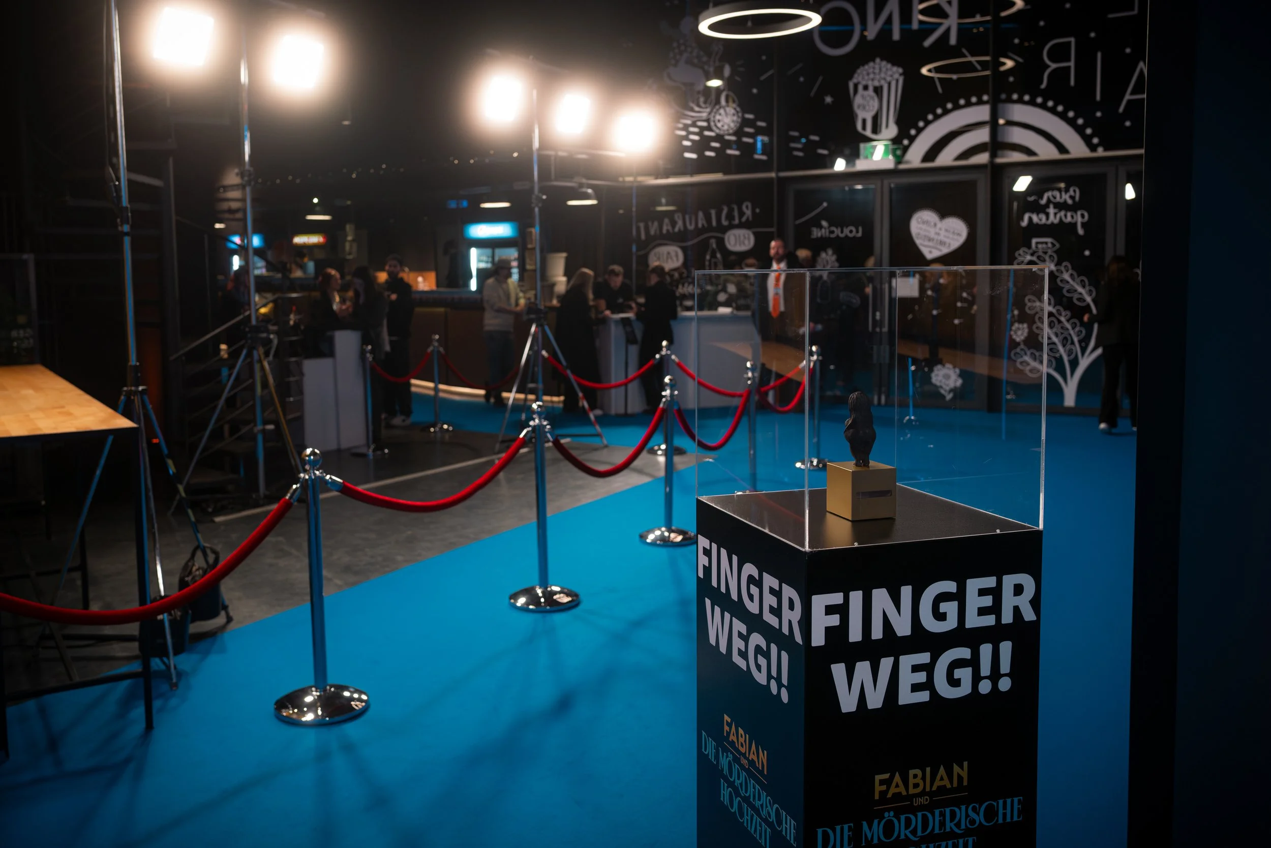 Ausstellung mit Skulptur unter einer Glasvitrine, roter Samtabsperrung, blaue Teppich, Menschen im Hintergrund in einem dunklen Raum mit Beleuchtung und decorativen Wandzeichnungen.