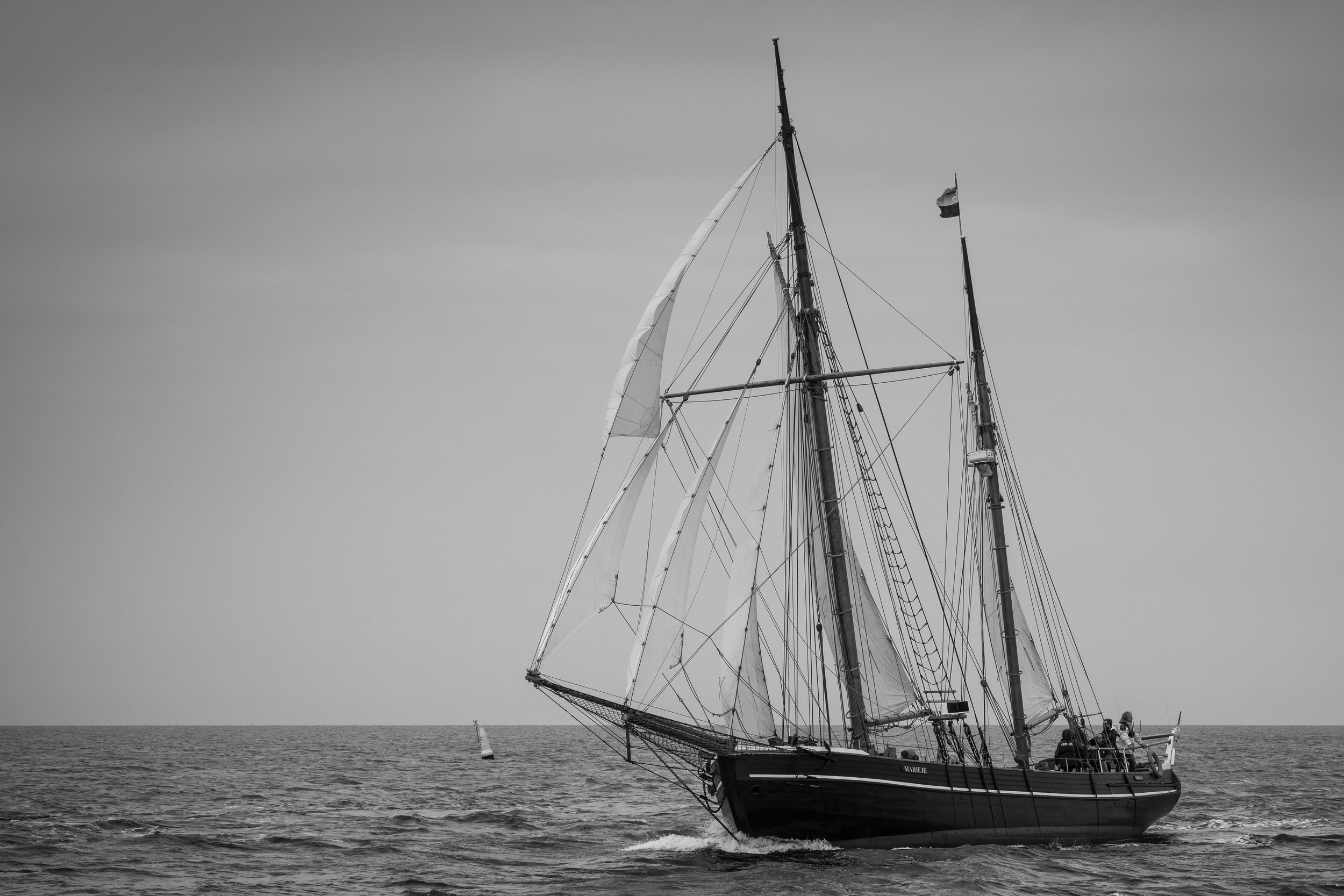 A black and white photograph of a sailing ship with raised sails on open water, with a small sailboat in the distance, under a cloudy sky.