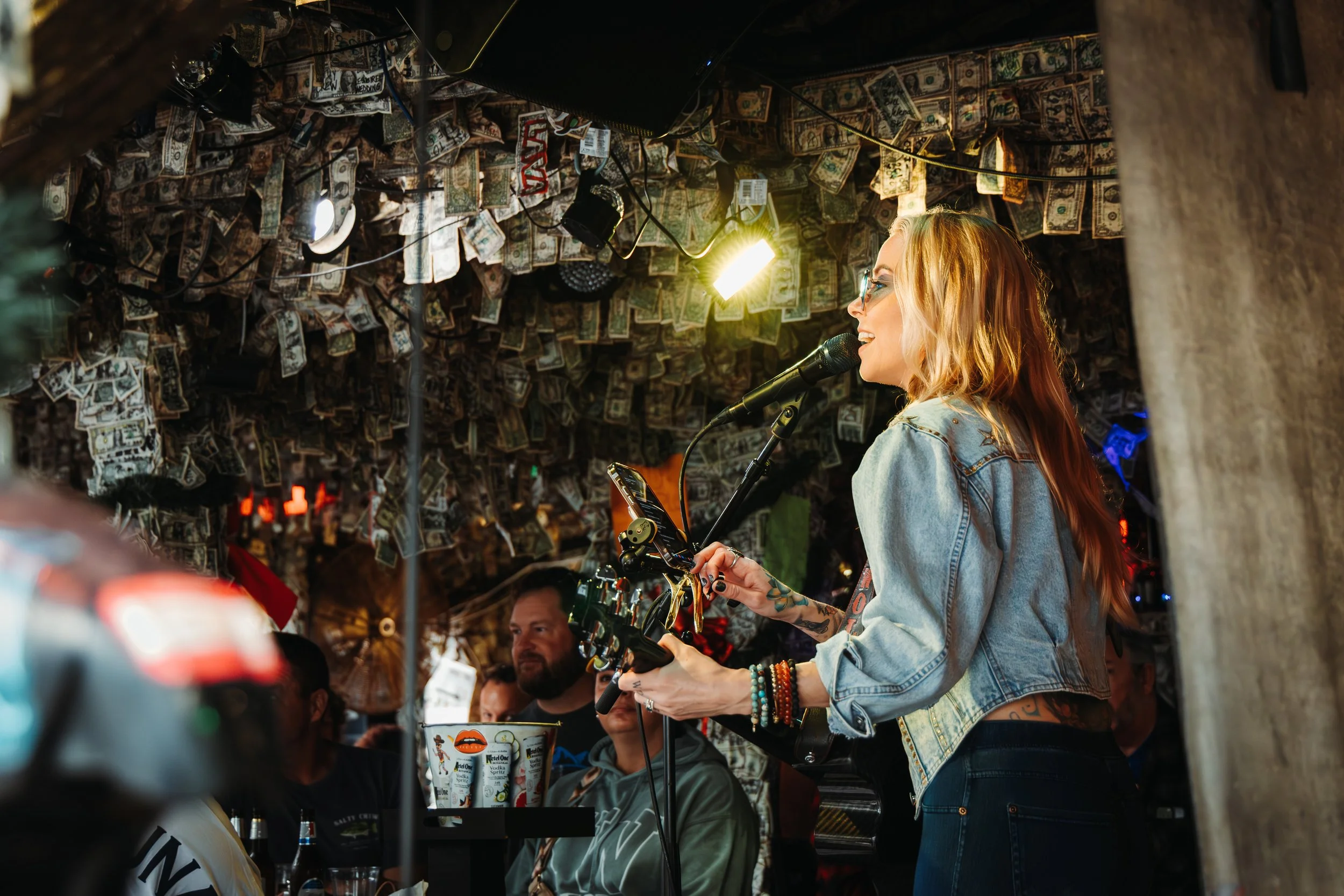 A woman with long red hair and tattoos, wearing a denim jacket, performs with a guitar and microphone in a bar decorated with dollar bills on the ceiling. Audience members are visible, watching her perform.