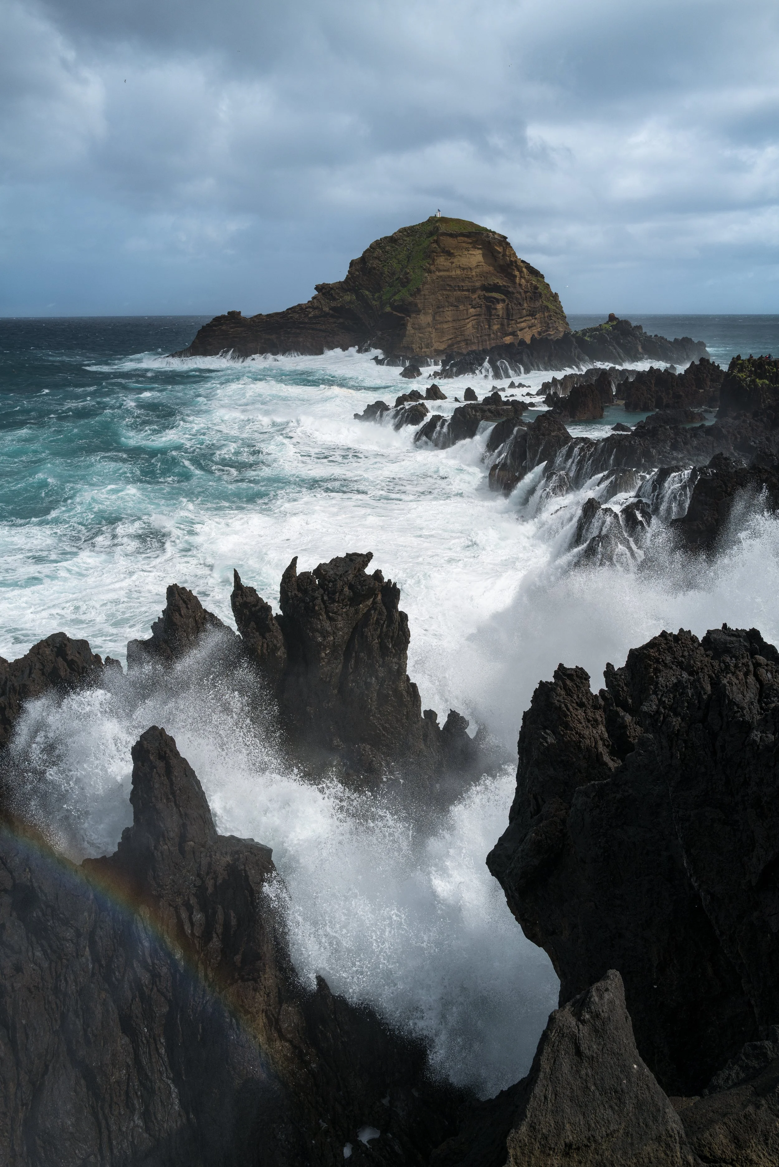 Rough ocean waves crashing against dark rocks with a small island in the background under a cloudy sky.