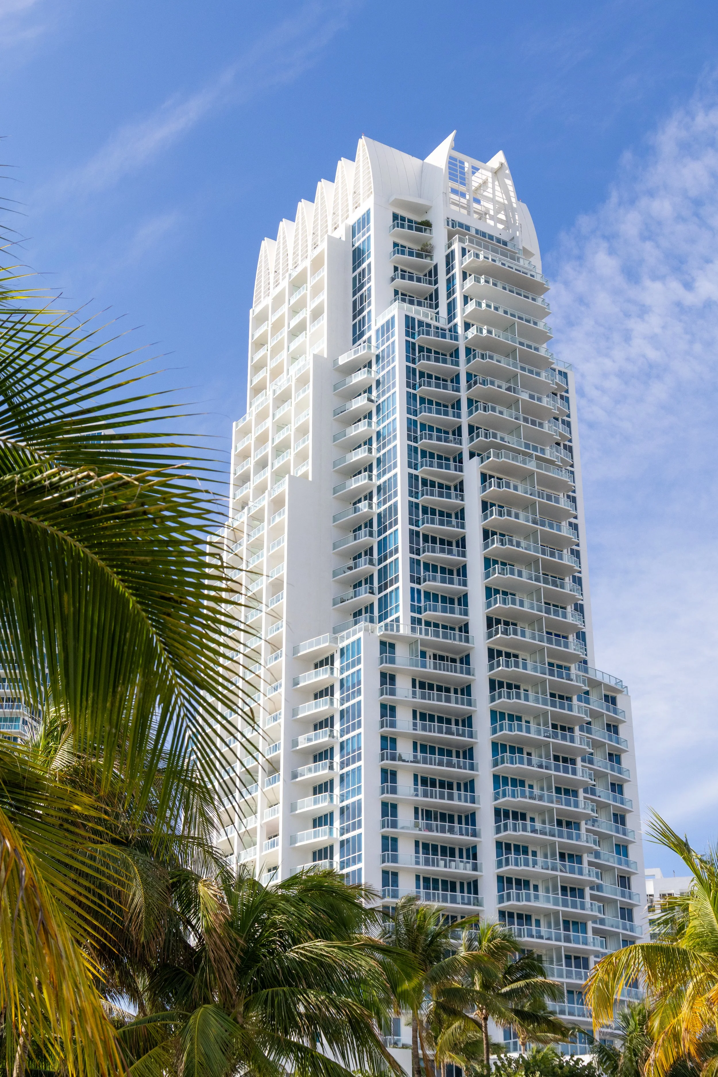 Tall modern white high-rise building with balconies, surrounded by palm trees under a blue sky.