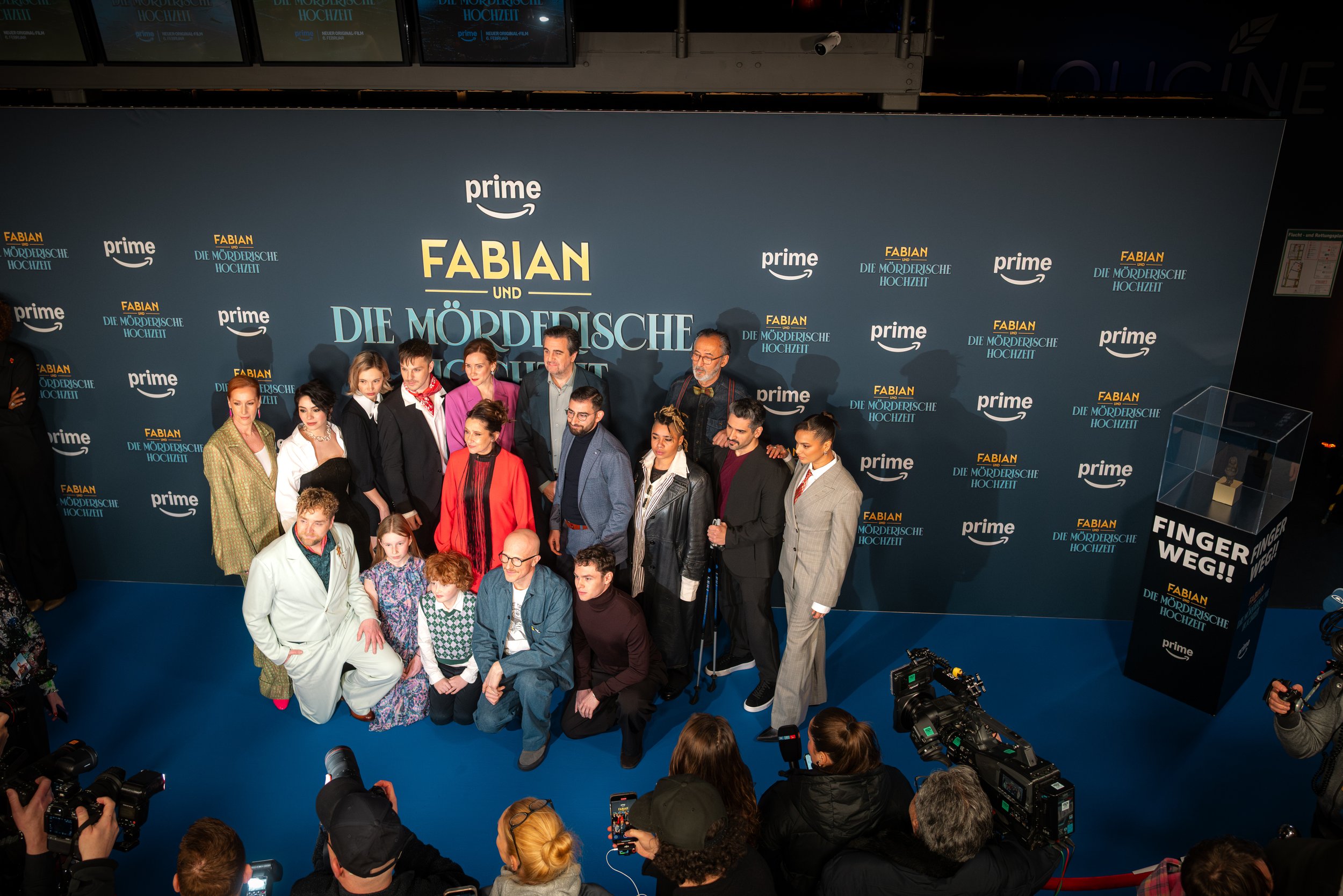 Gruppe von Darstellern und Filmmachern auf einem Roll-Up-Banner für die Fernsehserie 'Fabian und die mörderische Hochzeit' bei einer Presseveranstaltung, umgeben von Fotografen.