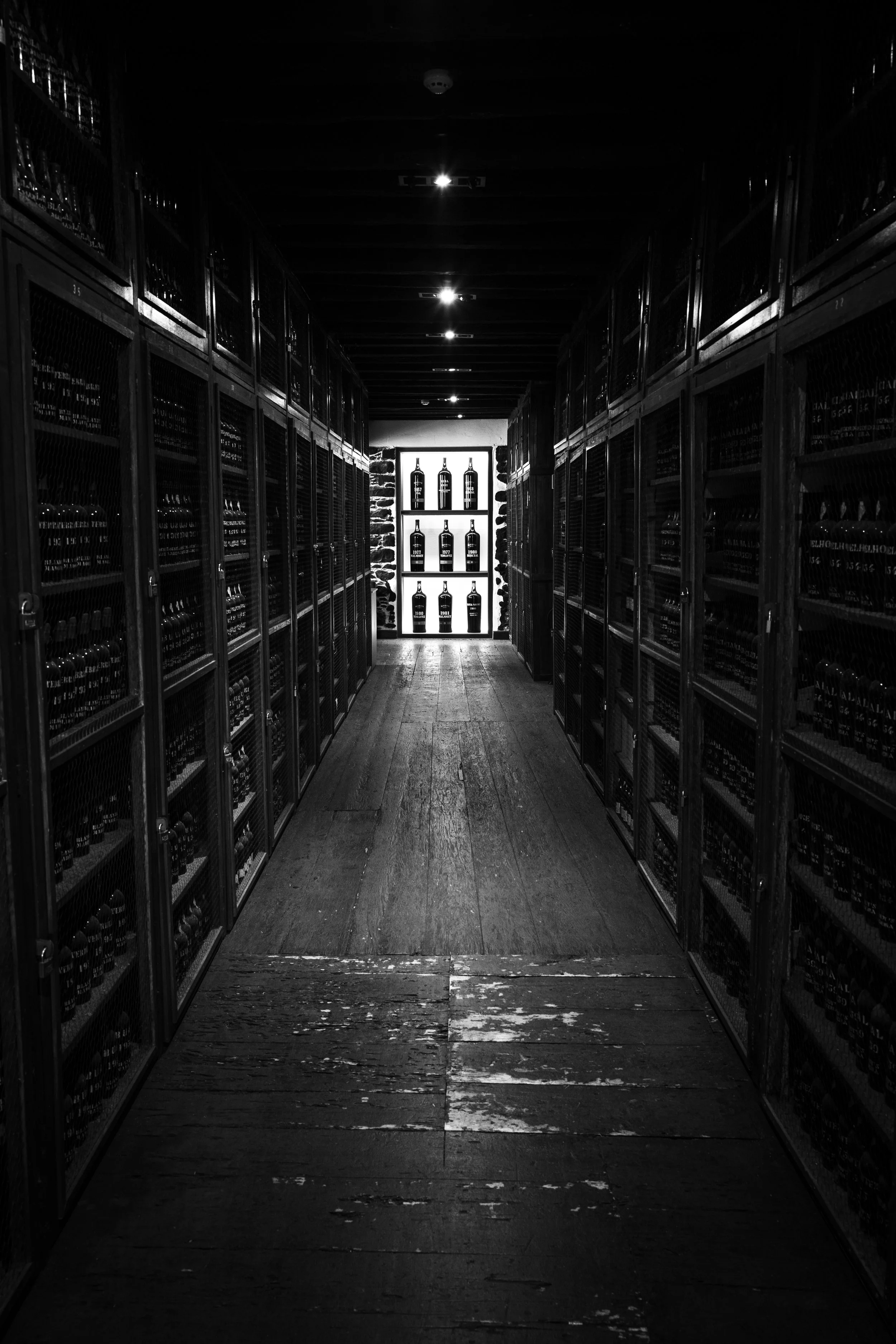 Black and white photo of a narrow wine cellar aisle with bottles stored on wooden shelves along both sides and illuminated bottles at the end of the aisle.