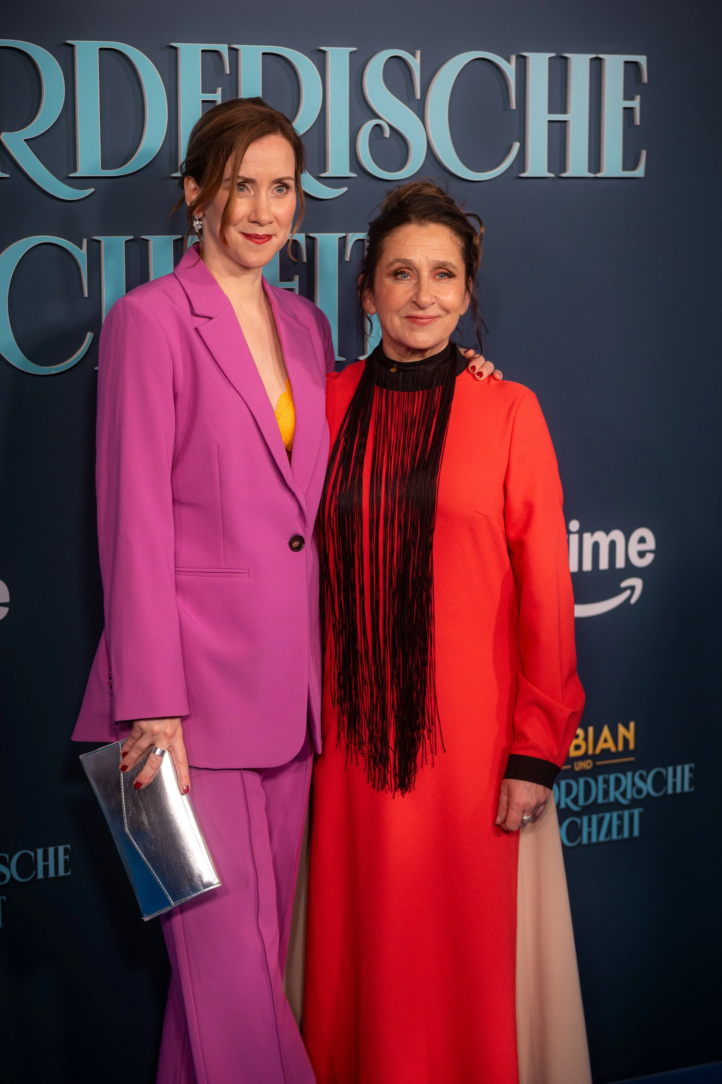 Zwei Frauen stehen nebeneinander vor einem blauen Hintergrund mit Text. Die Frau links trägt einen pinken Anzug und hält eine silberne Clutch, die Frau rechts trägt ein rotes Kleid mit schwarzen Fransen am Hals.