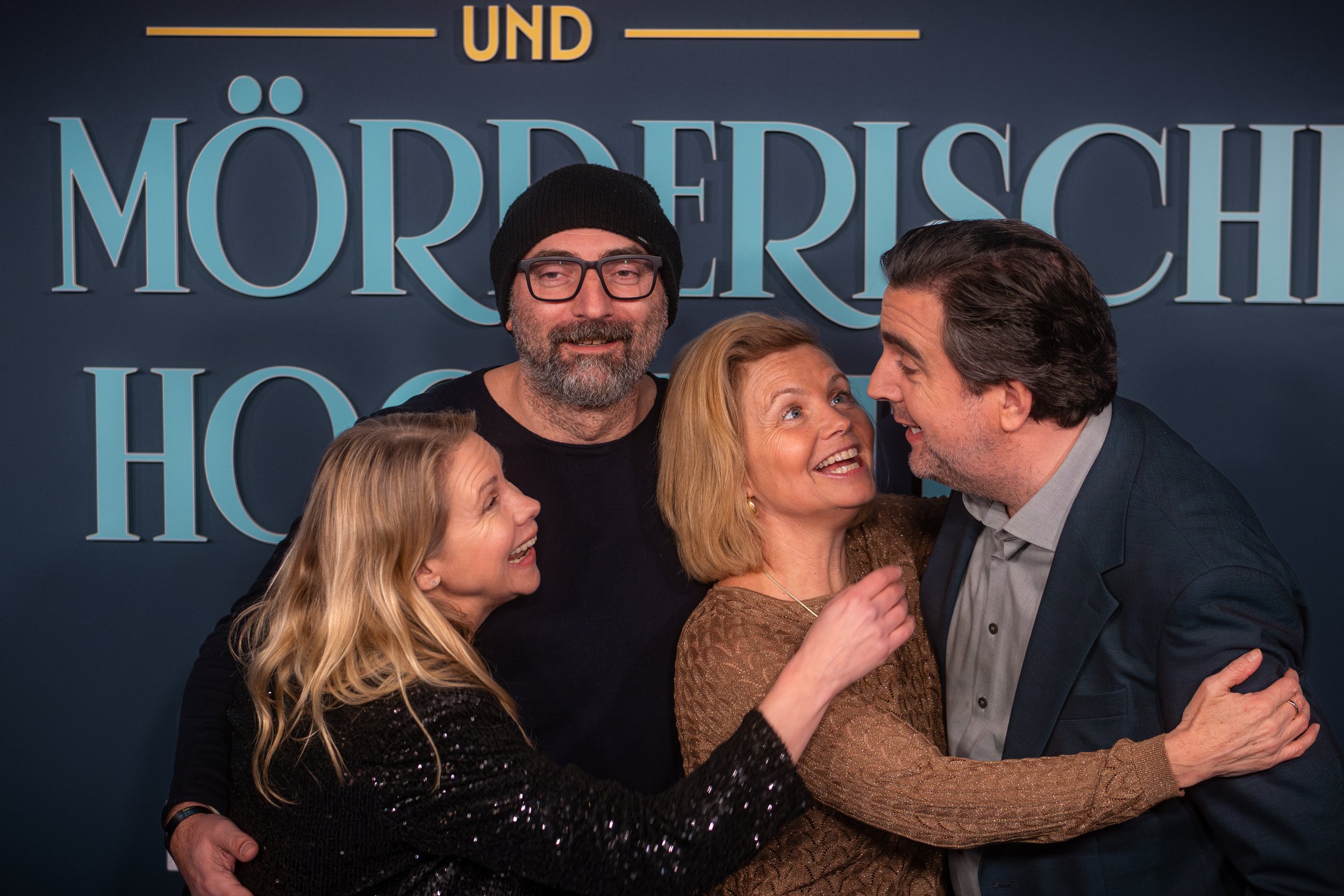 Gruppenfoto von vier lachenden Menschen, die sich umarmen und sich freudig begrüßen, vor einem blauen Hintergrund mit großen deutschen Schriftzügen.