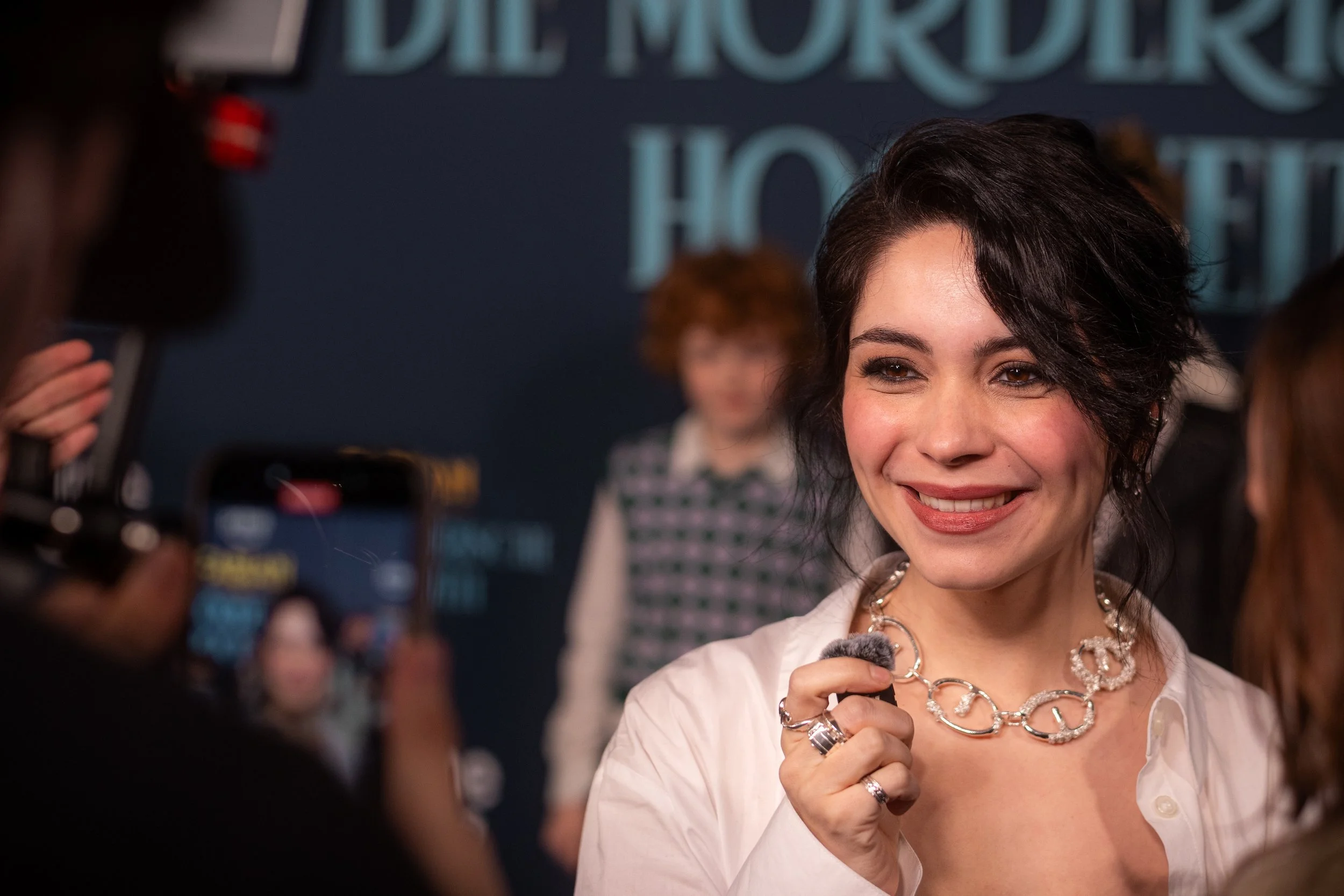 Eine lachende Frau mit dunklen, kurz gestylten Haaren trägt eine weiße Bluse und eine silberne, auffällige Halskette. Sie ist bei einem Interview oder Fotoshooting, im Hintergrund stehen andere Personen vor einer dunkelblauen Wand mit weißen Schriftz