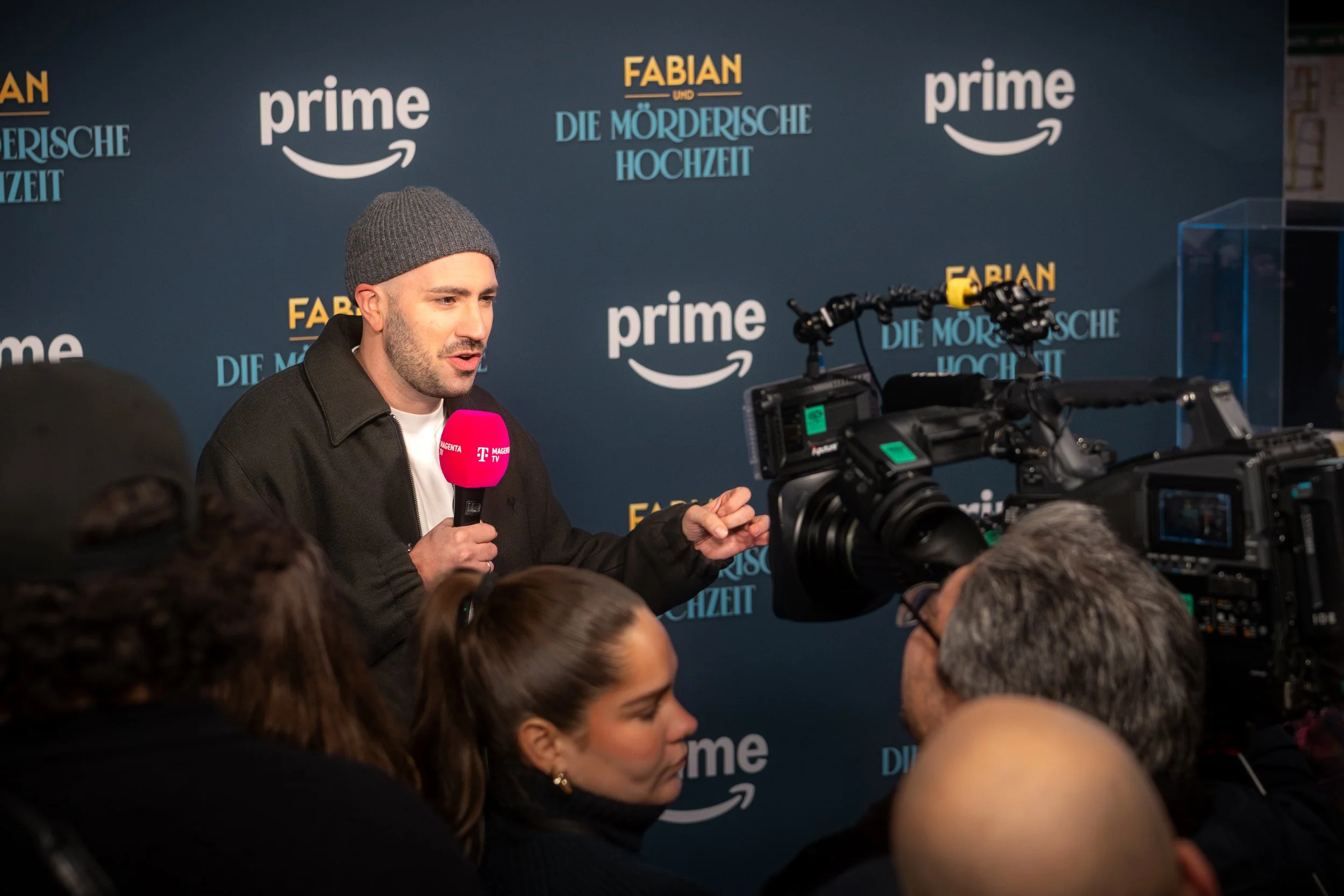 Mann mit grauem Beanie und schwarzen Jacke spricht in Mikrofon bei einer Veranstaltung, im Hintergrund Logos von Amazon Prime und 'Die mörderische Hochzeit'.