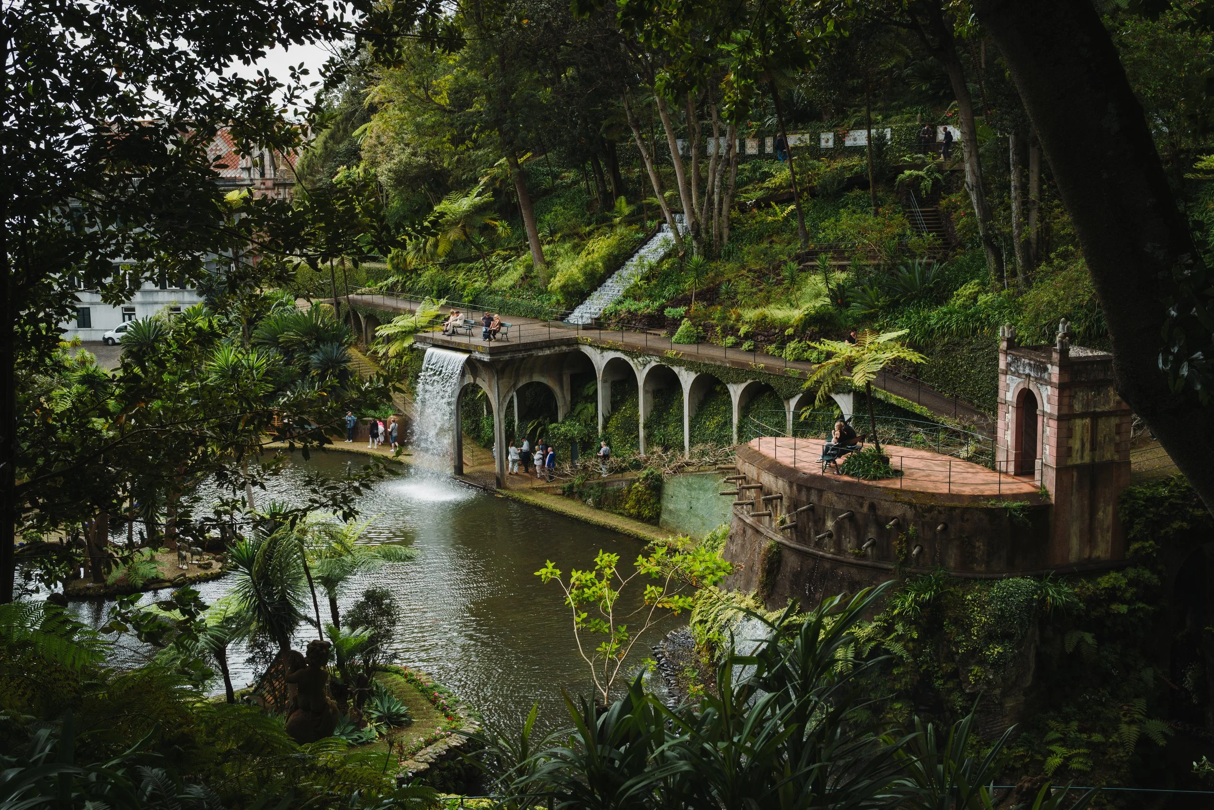 Lush green park with a river, waterfalls, and arched bridges. People are sitting on benches and walking along pathways, surrounded by trees and tropical plants.