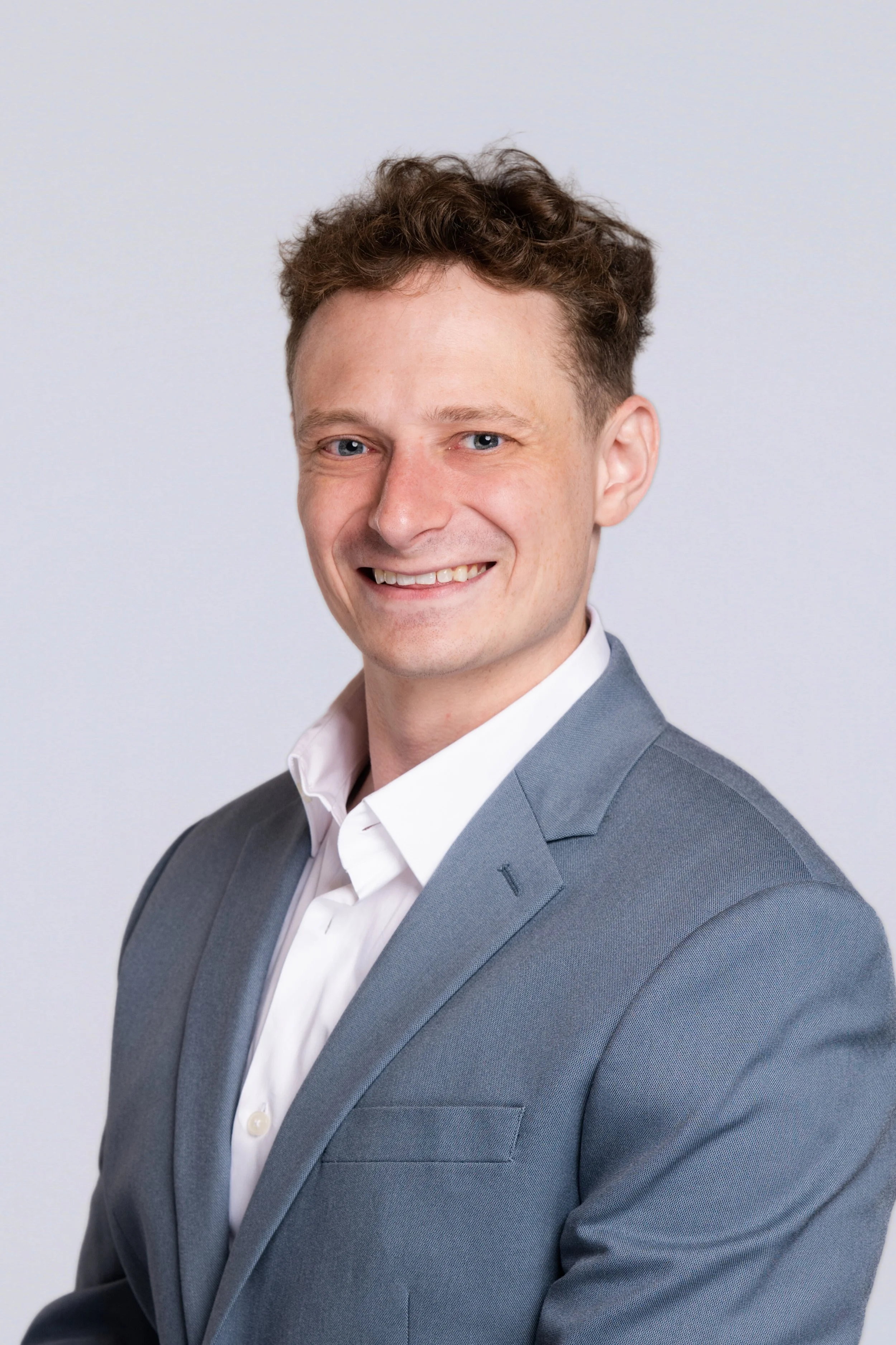 Portrait of a smiling man with curly brown hair wearing a grey suit and white shirt against a plain light background.