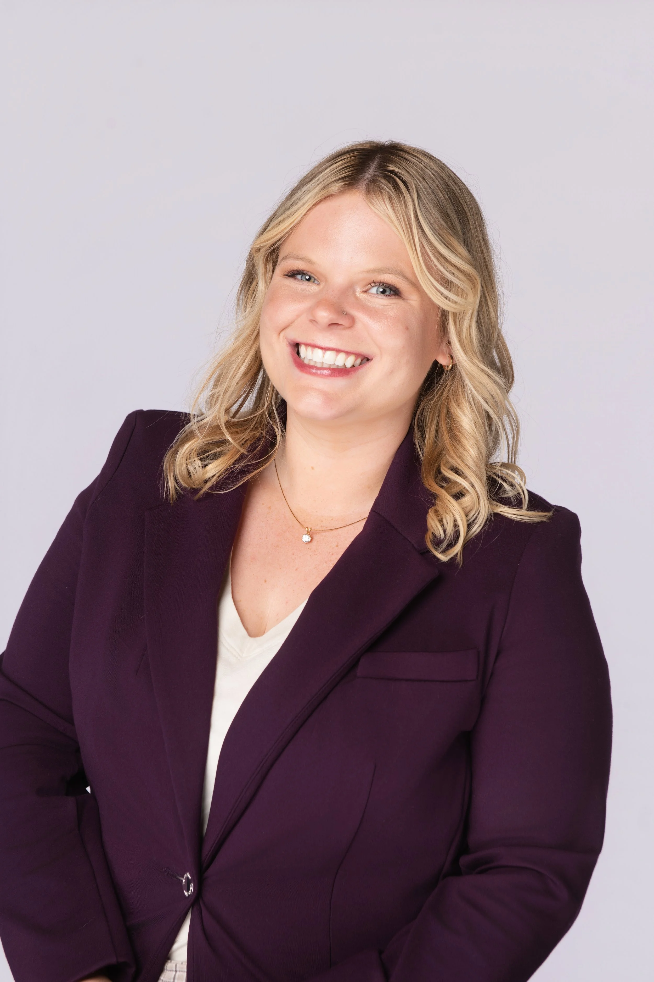 A young woman with blonde wavy hair is smiling and wearing a dark purple blazer over a white top, standing against a plain white background.