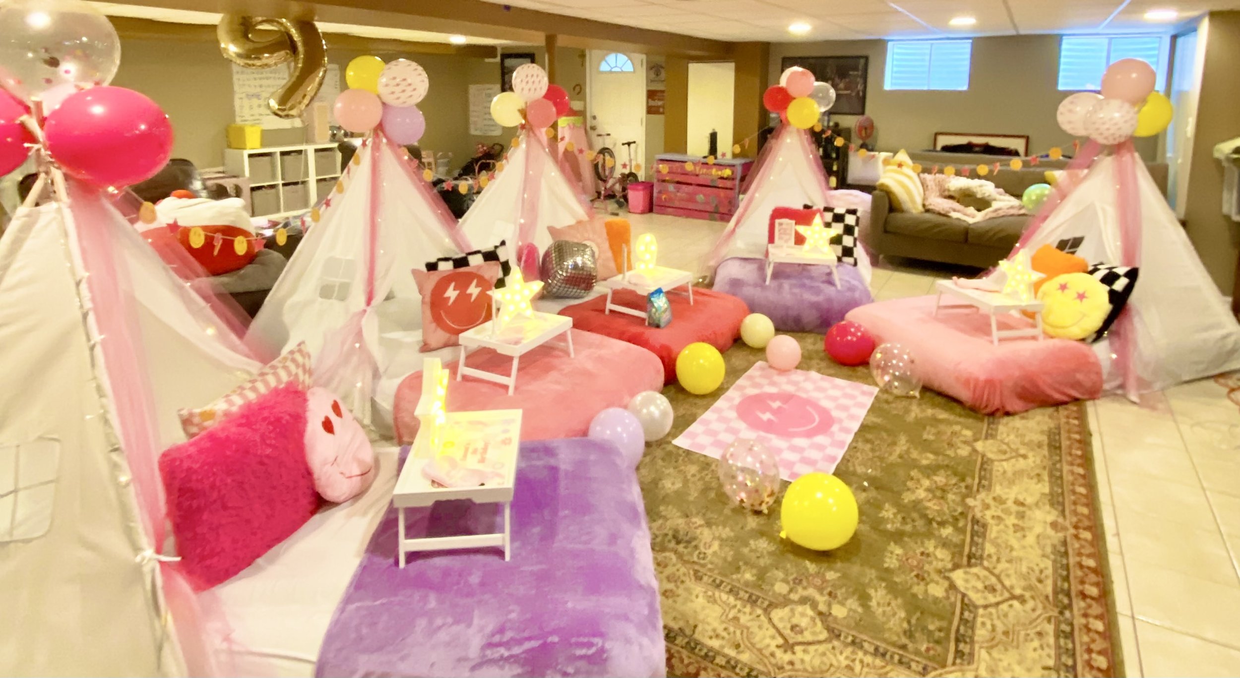Colorful indoor birthday party setup with pink, purple, yellow, and white balloons, small tents, pillows, and fairy lights in a basement room.