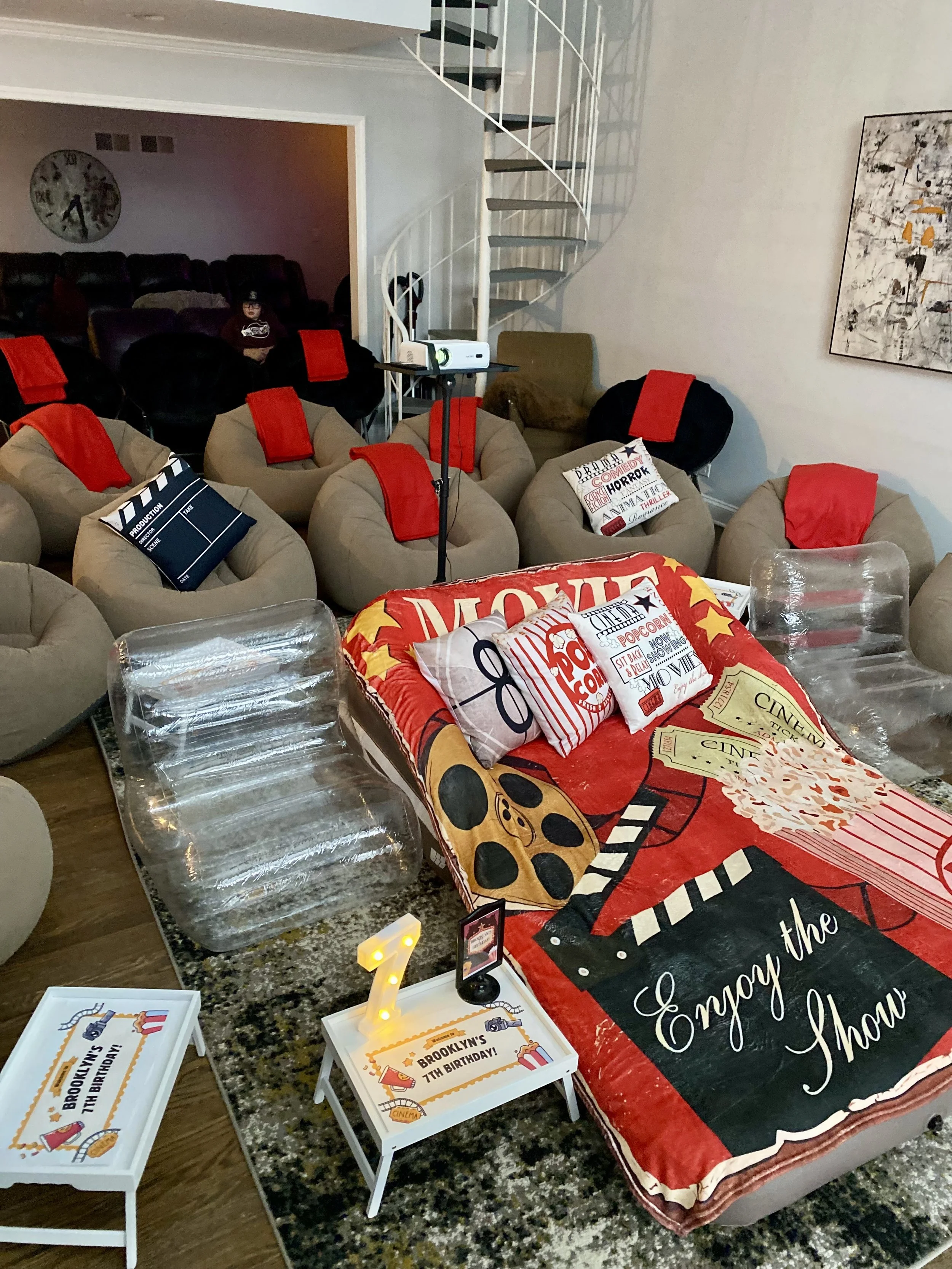Living room decorated for a birthday party with a movie theme, featuring a large popcorn and movie reel themed blanket, plush chairs with red towels, and a TV featuring movie-related pillows and banners.