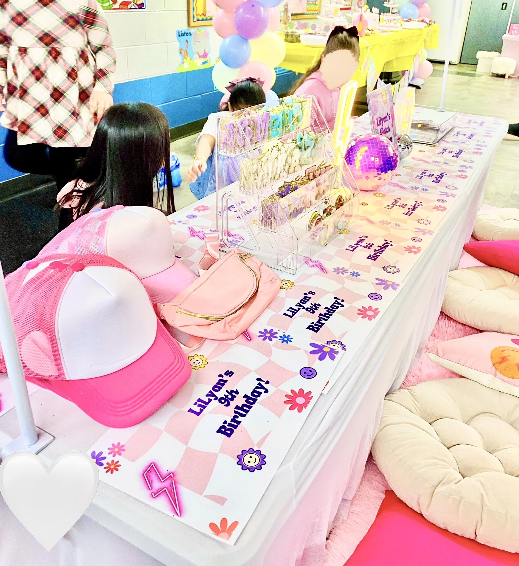 A decorated birthday party table with pink and purple theme, featuring three pink and white hats, a pink bag, various party favors, and balloons in pastel colors. Kids are gathered around the table.