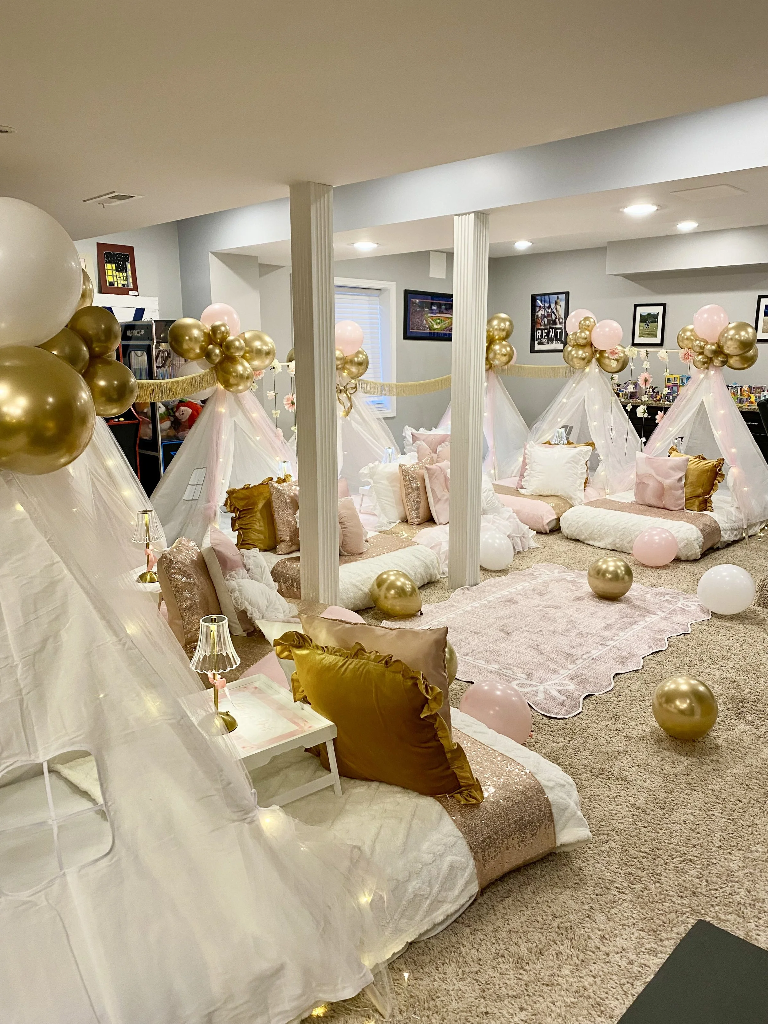 Table decorated with pink, gray, and white balloons, with decor including books, neon flamingo lights, small boxes, and engraved cards, set against a windowed background with sunlight and a swimming pool visible outside.