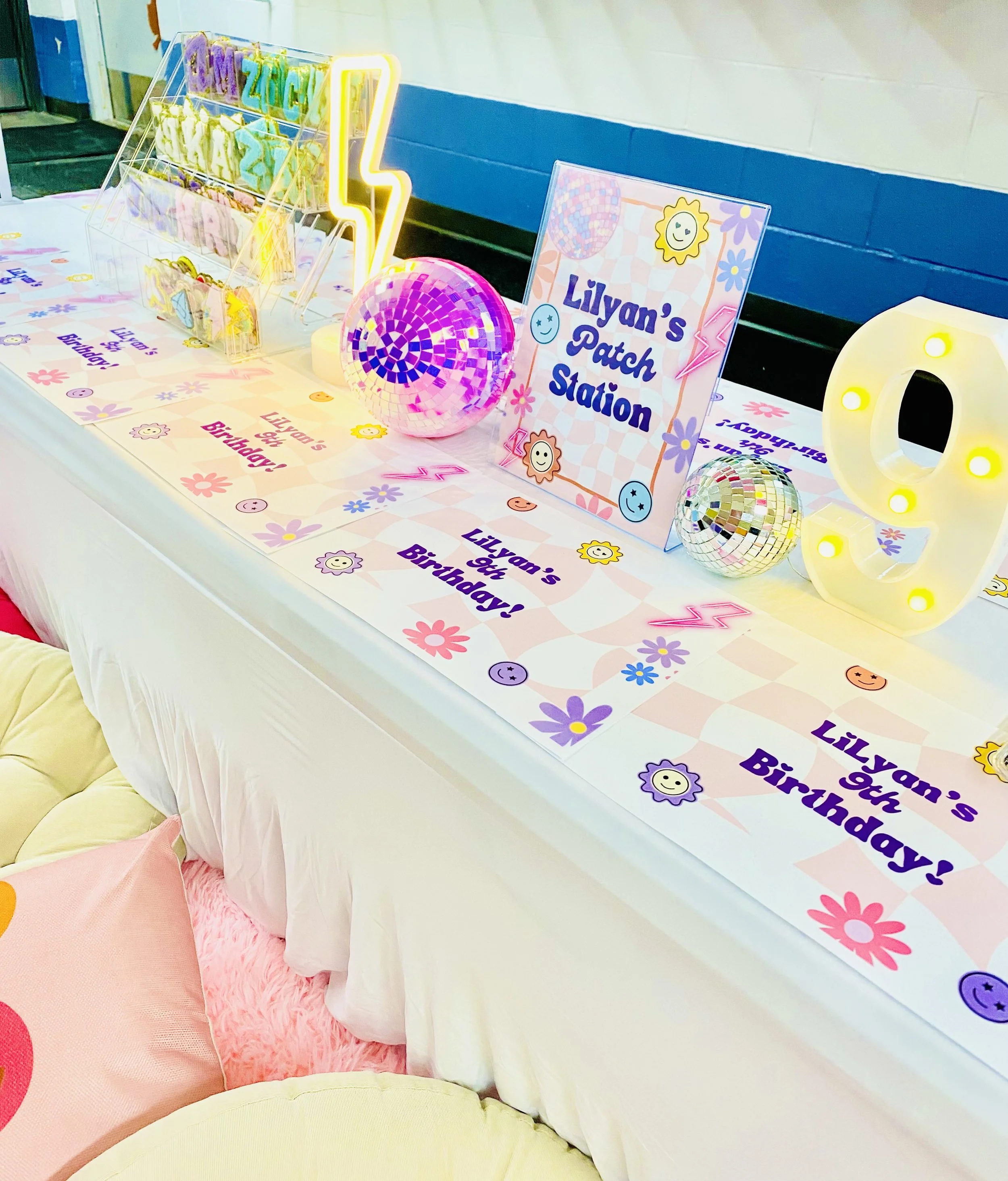 Decorative birthday party table with a sign reading 'Lilyan's Patch Station,' colorful disco balls, a large illuminated letter 'Q,' and a sign with the same text. The table features pastel-colored flowers and smiley face designs, with a pink, yellow, and white theme.
