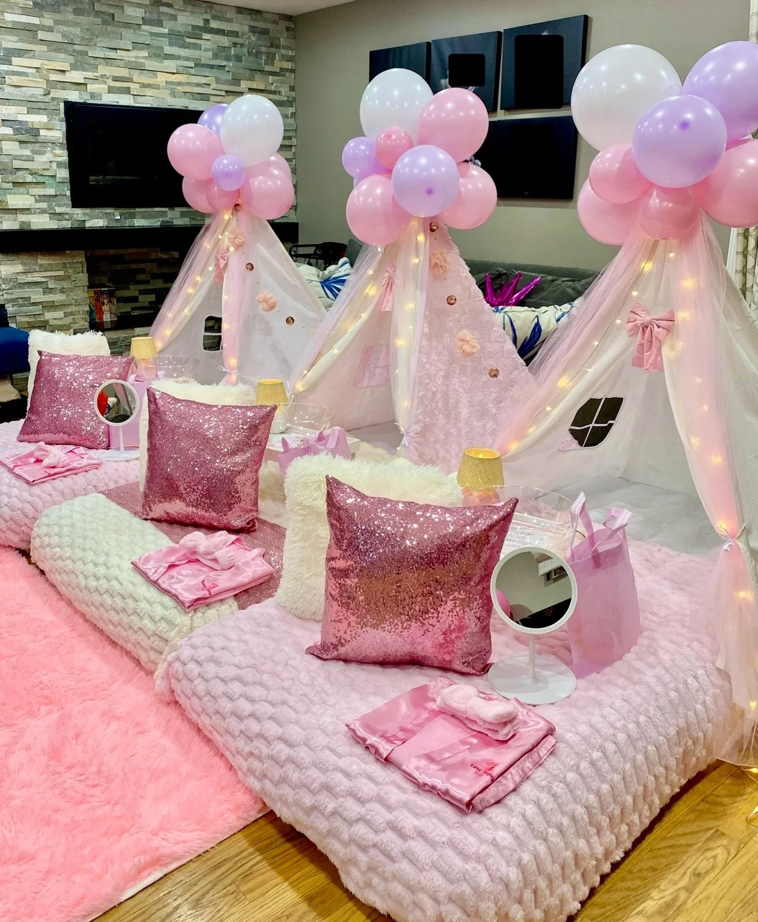 A cozy indoor space decorated for a girls' sleepover or party, featuring pink and purple balloons and tents with fairy lights, pink pillows, and pink satin gift bags on a pink blanket.