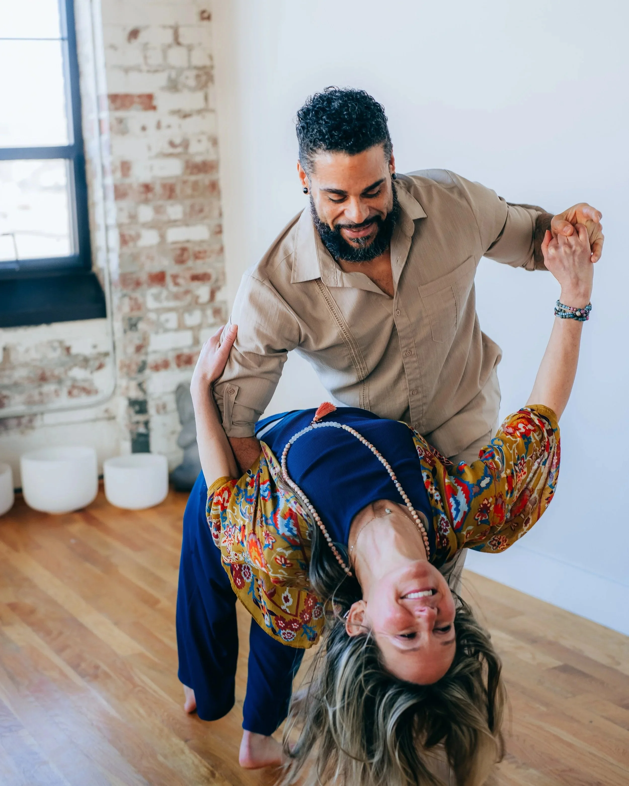 A person in a colorful jacket and dark pants is performing a yoga pose with their upper body leaning back and their head upside down, supported by someone else. The background features a brick wall, large window, and wooden floor.