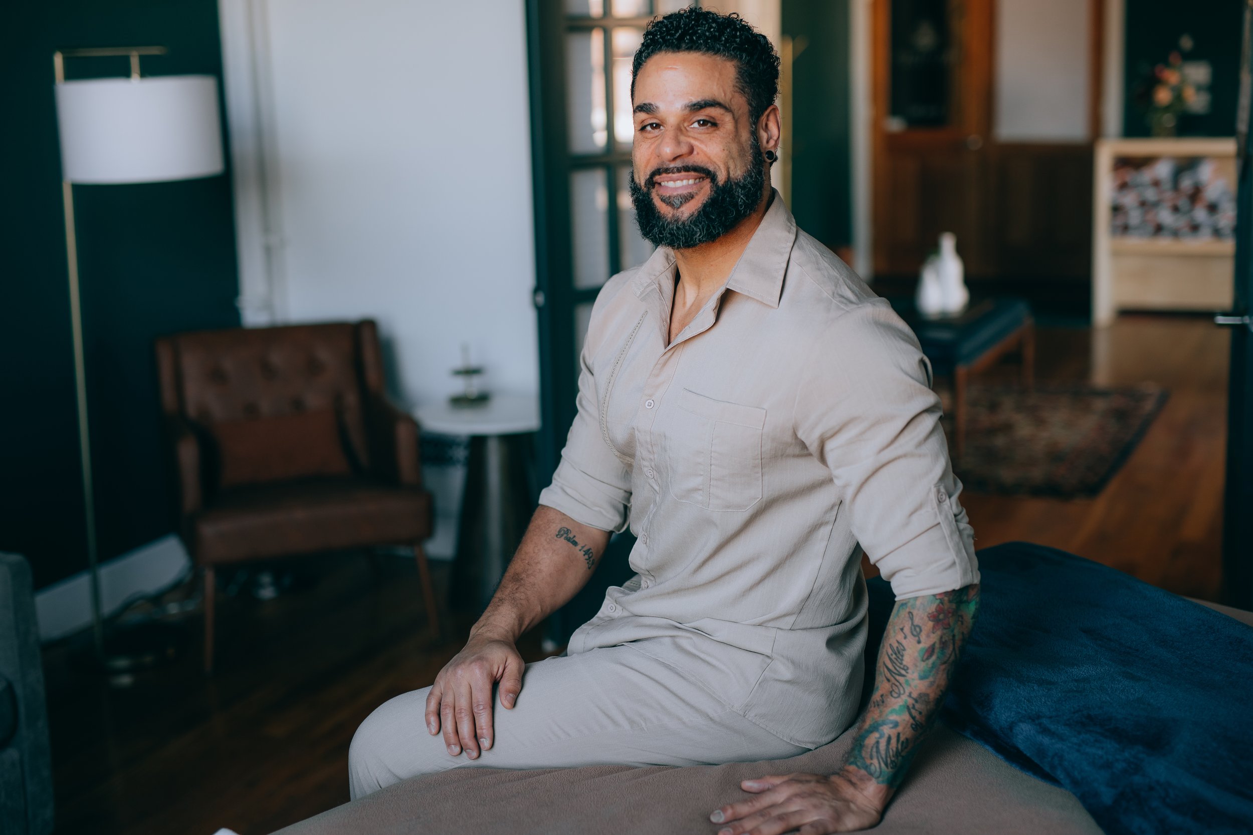 A man with curly hair and a beard sitting on a bed in a modern room, smiling at the camera with a tattooed arm.
