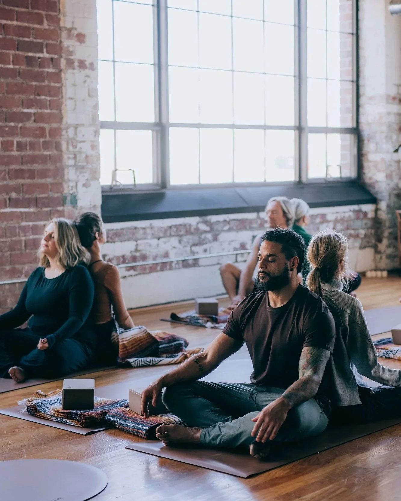 People practicing yoga in a spacious, industrial-style room with exposed brick walls and large windows, sitting cross-legged on yoga mats.