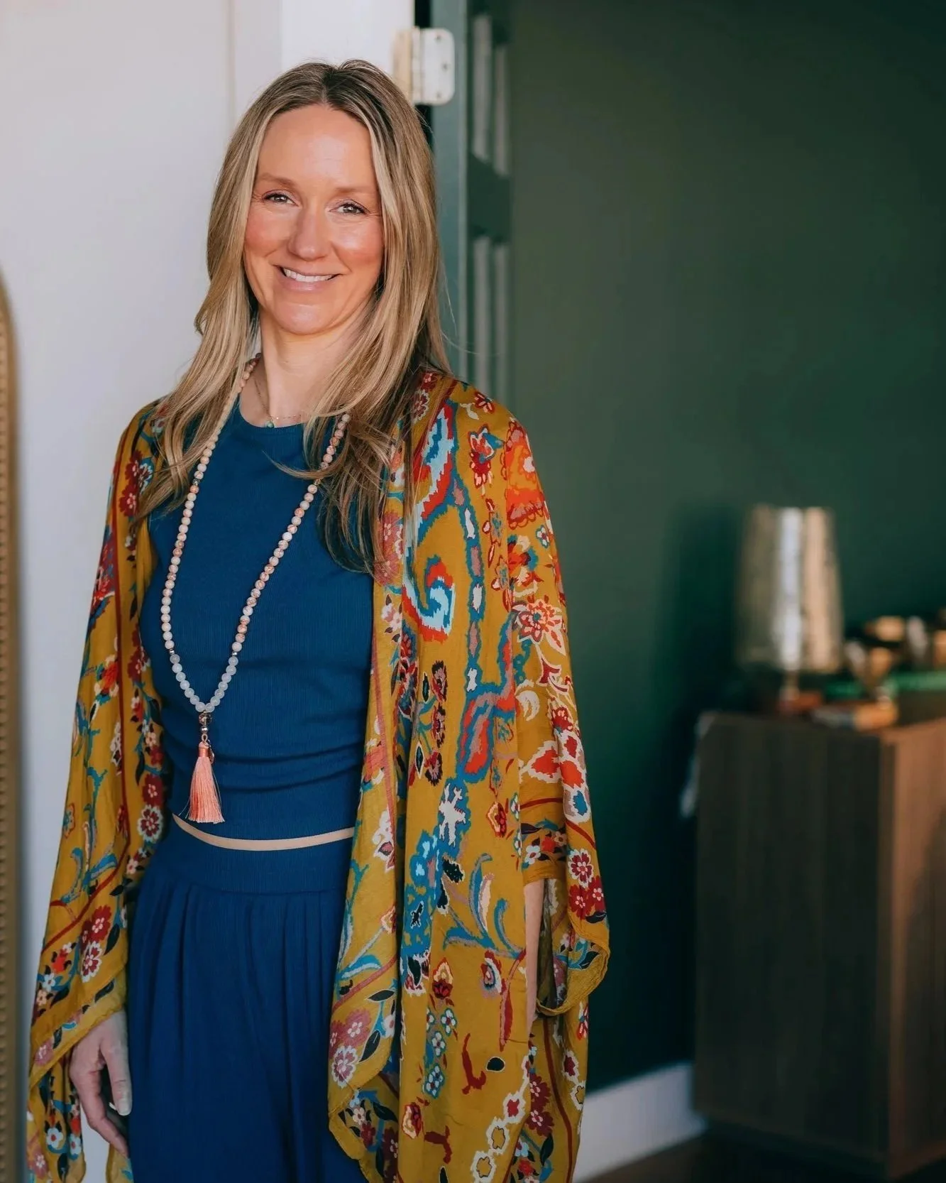 A woman with long blonde hair smiling, wearing a colorful patterned kimono over a blue dress, standing indoors with a dark green wall and blurred objects in the background.