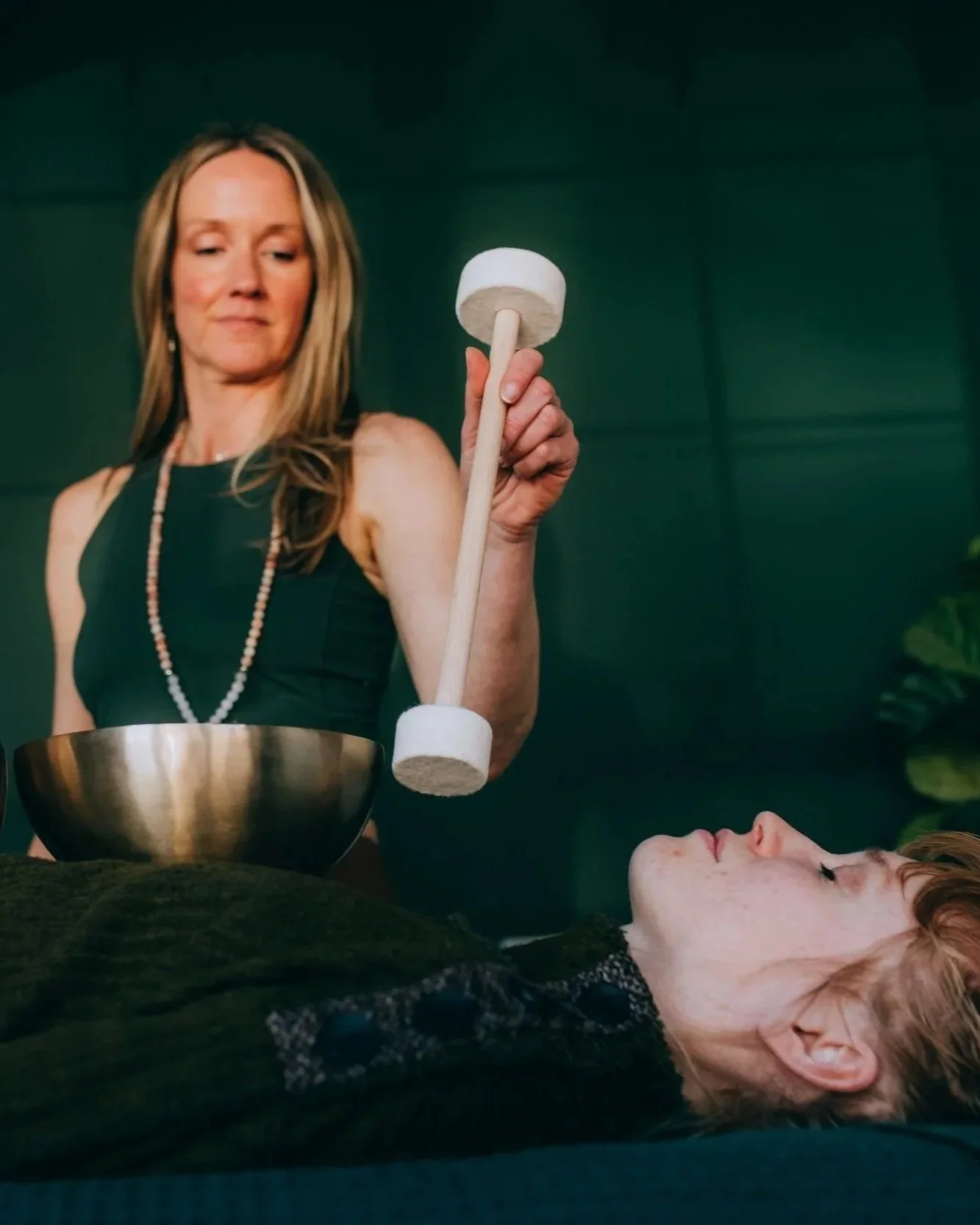A woman with long blonde hair is holding a large mallet above another woman lying on a table. The woman on the table has red hair and is resting with eyes closed, wearing a black lace top, in a room with dark green walls.