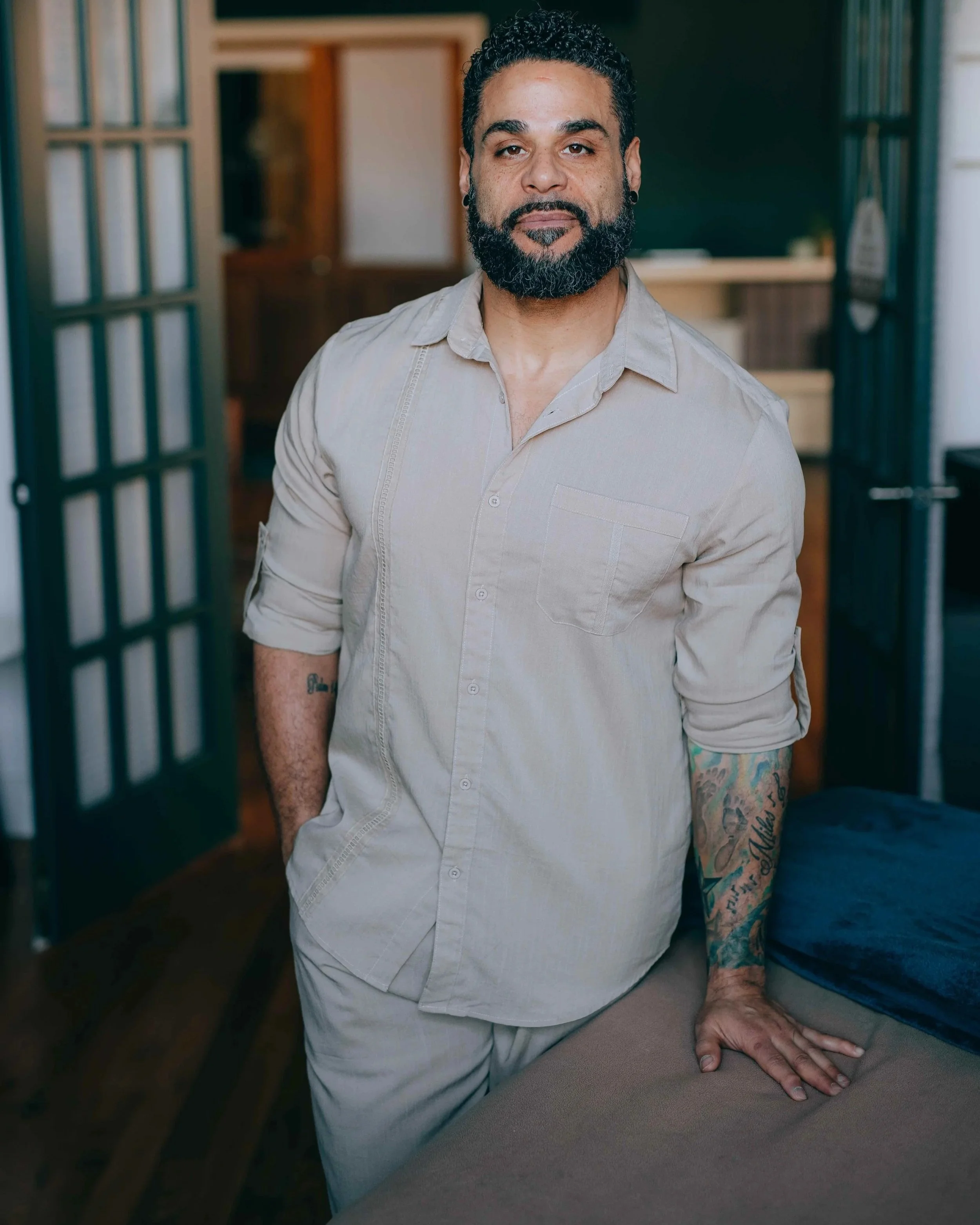 A man with a beard and tattoos wearing a light-colored shirt with sleeves rolled up, leaning on a table with his hand resting on it, in an indoor setting with wooden furniture and a chalkboard in the background.
