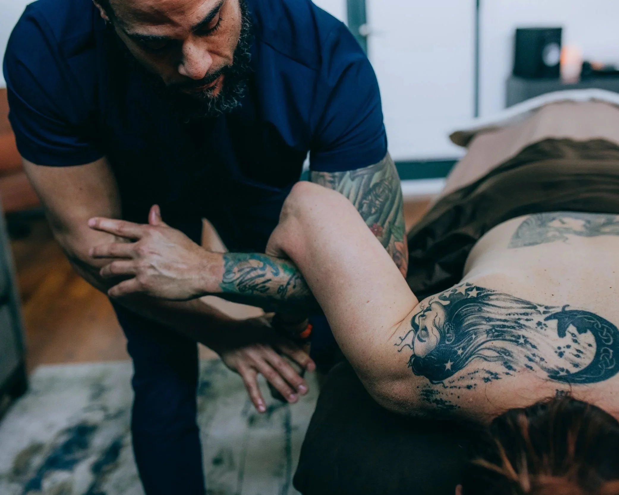 A man with a beard and tattoos on his arms leaning over a person's arm, appearing to perform a tattoo or similar activity in a room with a bed and a window.